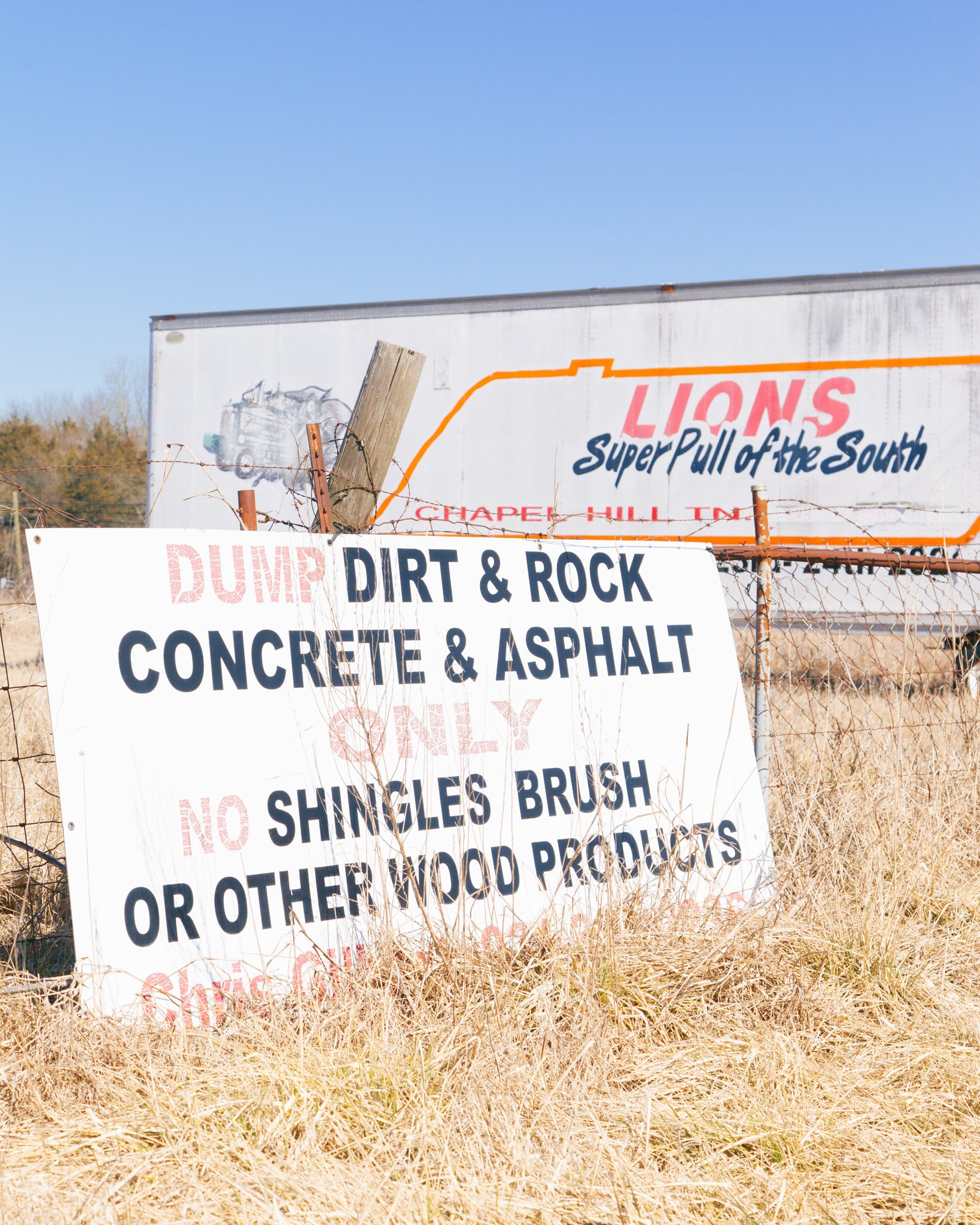 Close-up of a weathered outdoor sign on a wire fence with dry grass in the foreground and a clear blue sky in the background. The sign advertises dirt and rock disposal services and mentions specific types of materials accepted.