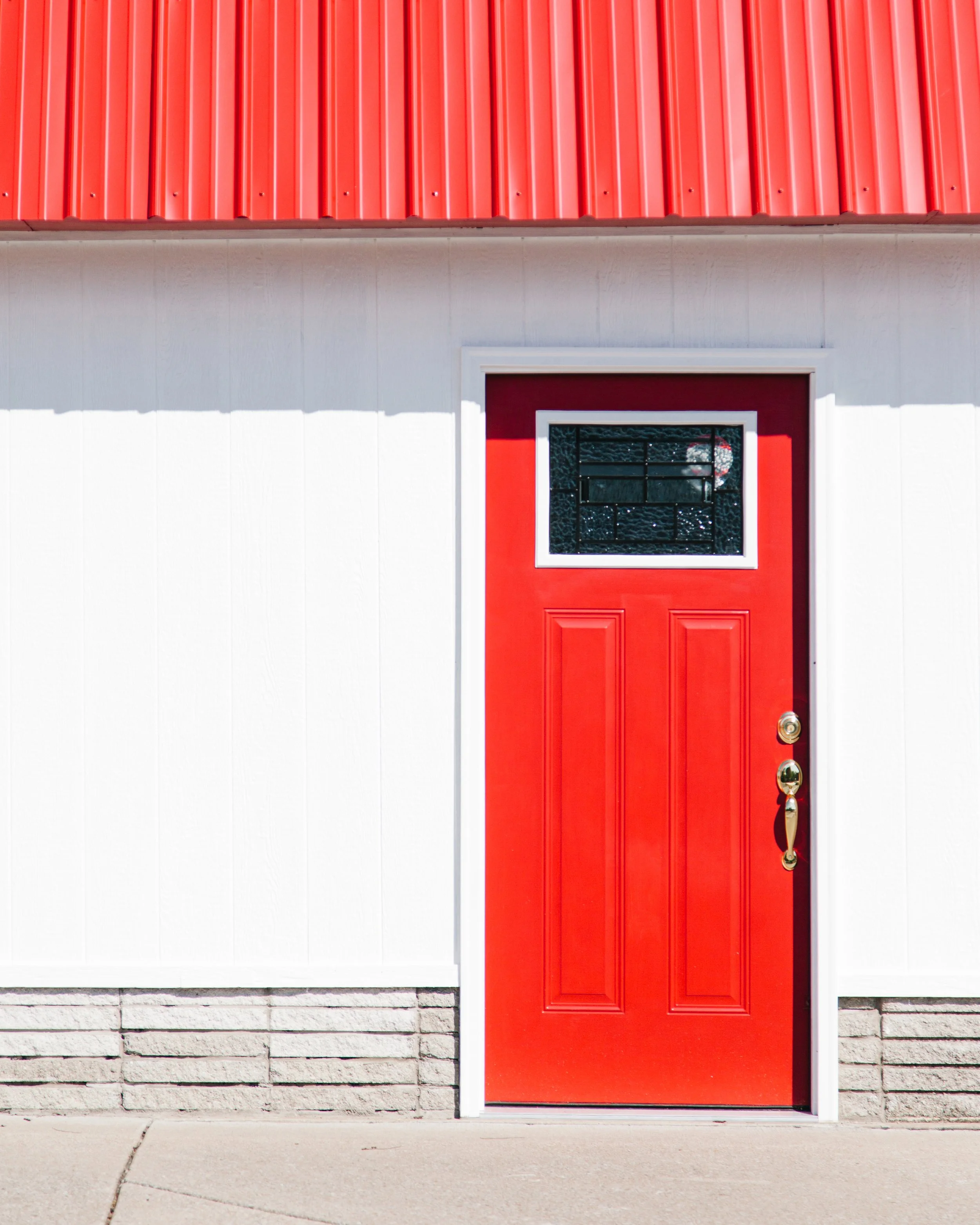 A bright red door with a small window, set in a white building with brick foundation, viewed from the sidewalk.