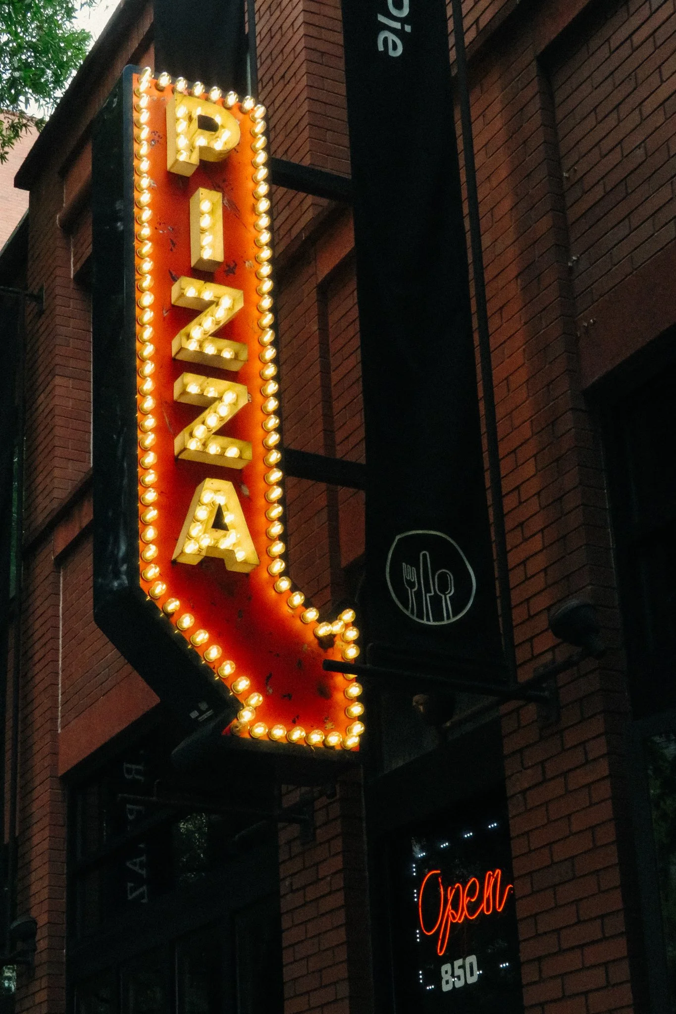 Neon sign with the word 'PIZZA' outlined in small light bulbs hanging on a brick building exterior. Another sign below reads 'Open' in red neon and has some additional text and numbers.