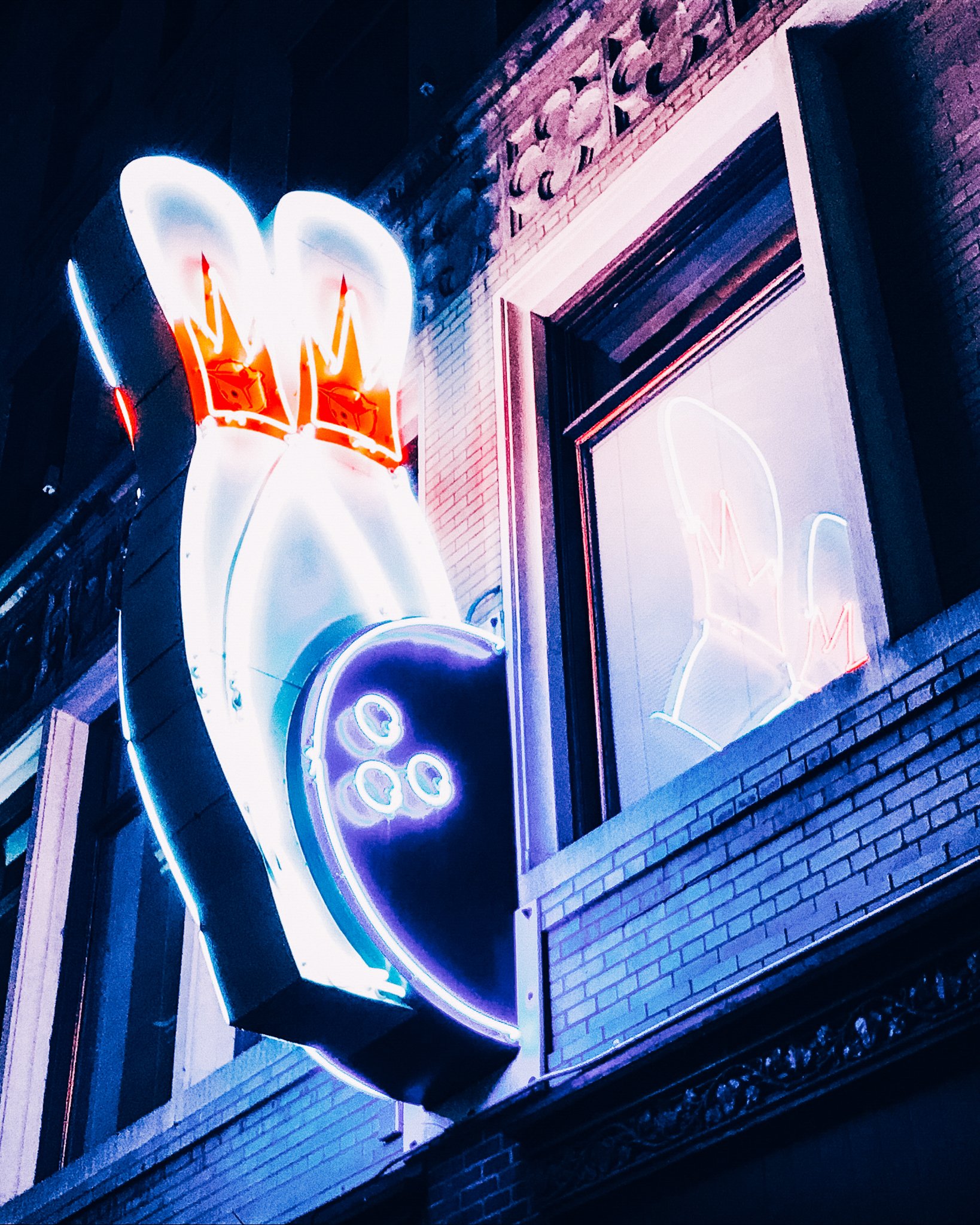 Colorful neon sign on a brick building at night, featuring a stylized image of a soda cup with a straw. The sign is illuminated with bright neon lights in red, blue, and white, creating a vibrant glow.