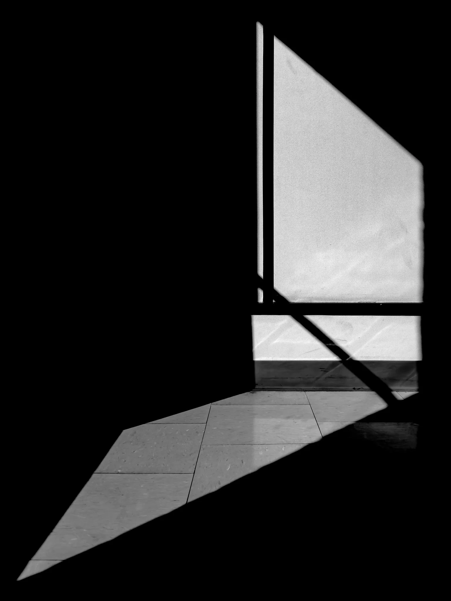 A black and white photo of a door with a window, partially open, with sunlight casting shadows on the floor and wall. The photograph is taken from inside a dark room.