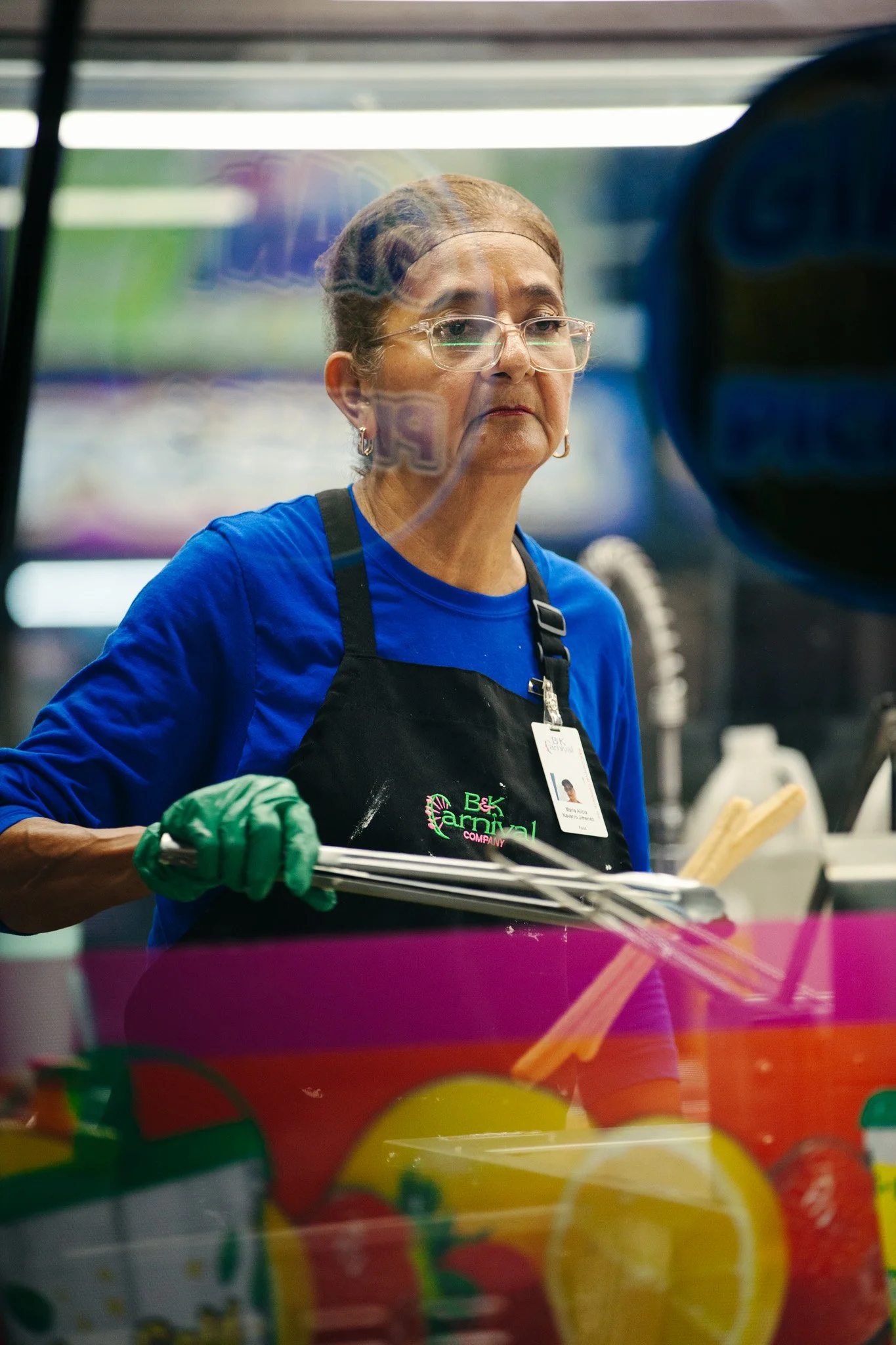 A woman wearing glasses, a blue shirt, and a black apron with colorful text is seen through a glass window, possibly working at a food or beverage stand, with various colorful items in the foreground.