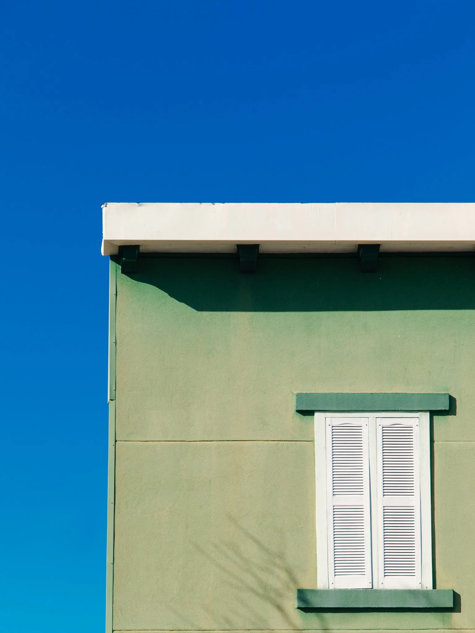 A green building with white shutters on a window, against a clear bright blue sky.
