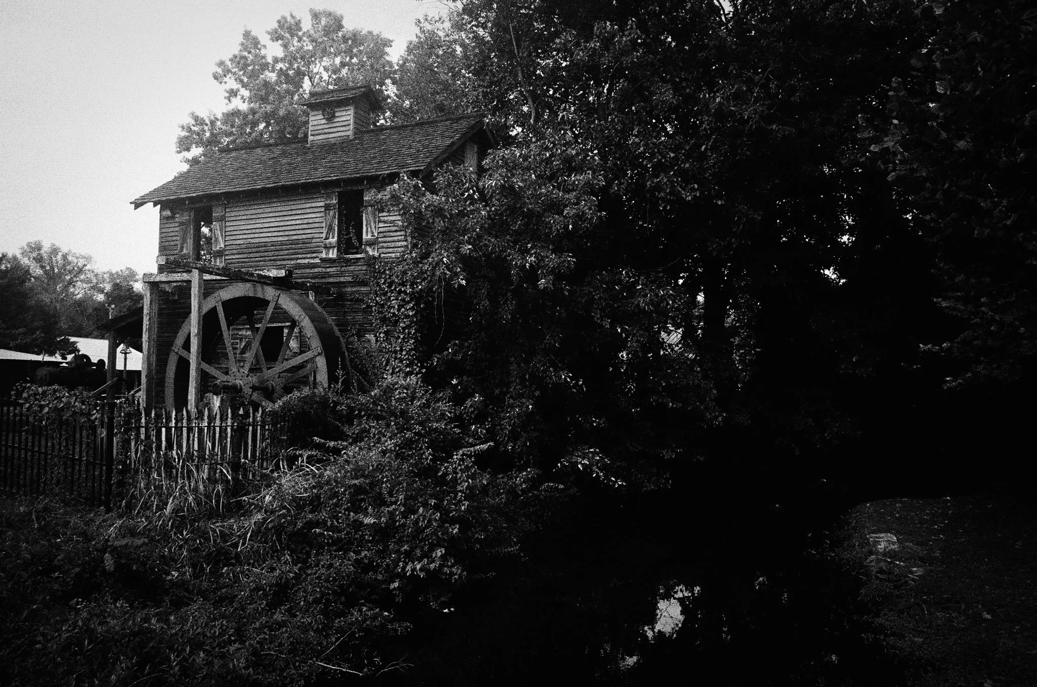 Black and white photo of an old wooden watermill with a large wheel, surrounded by trees and a fence in the foreground.