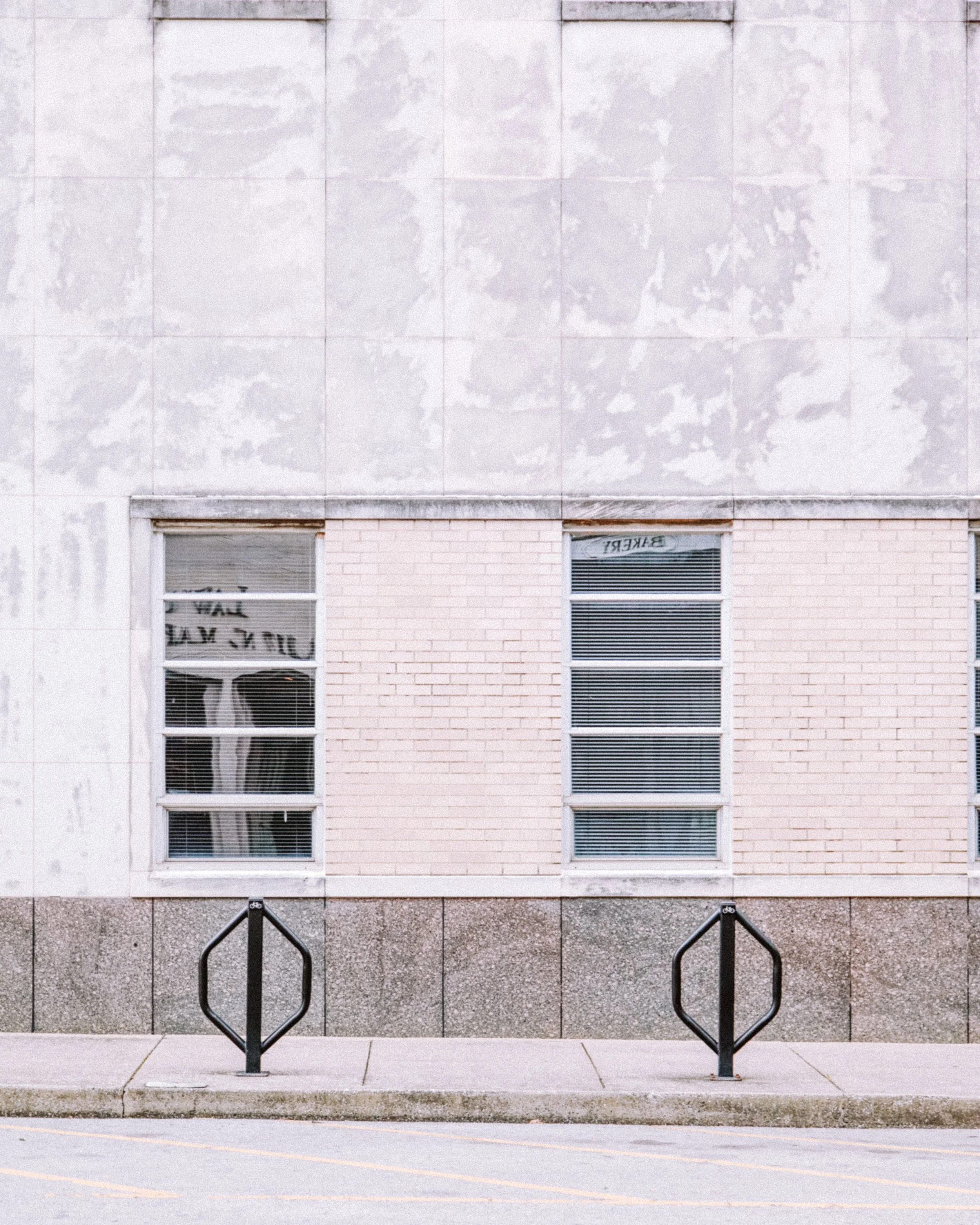 A brick and concrete building with three large windows and two black bicycle racks in front, along with a sidewalk and a parking lot marked by yellow lines.