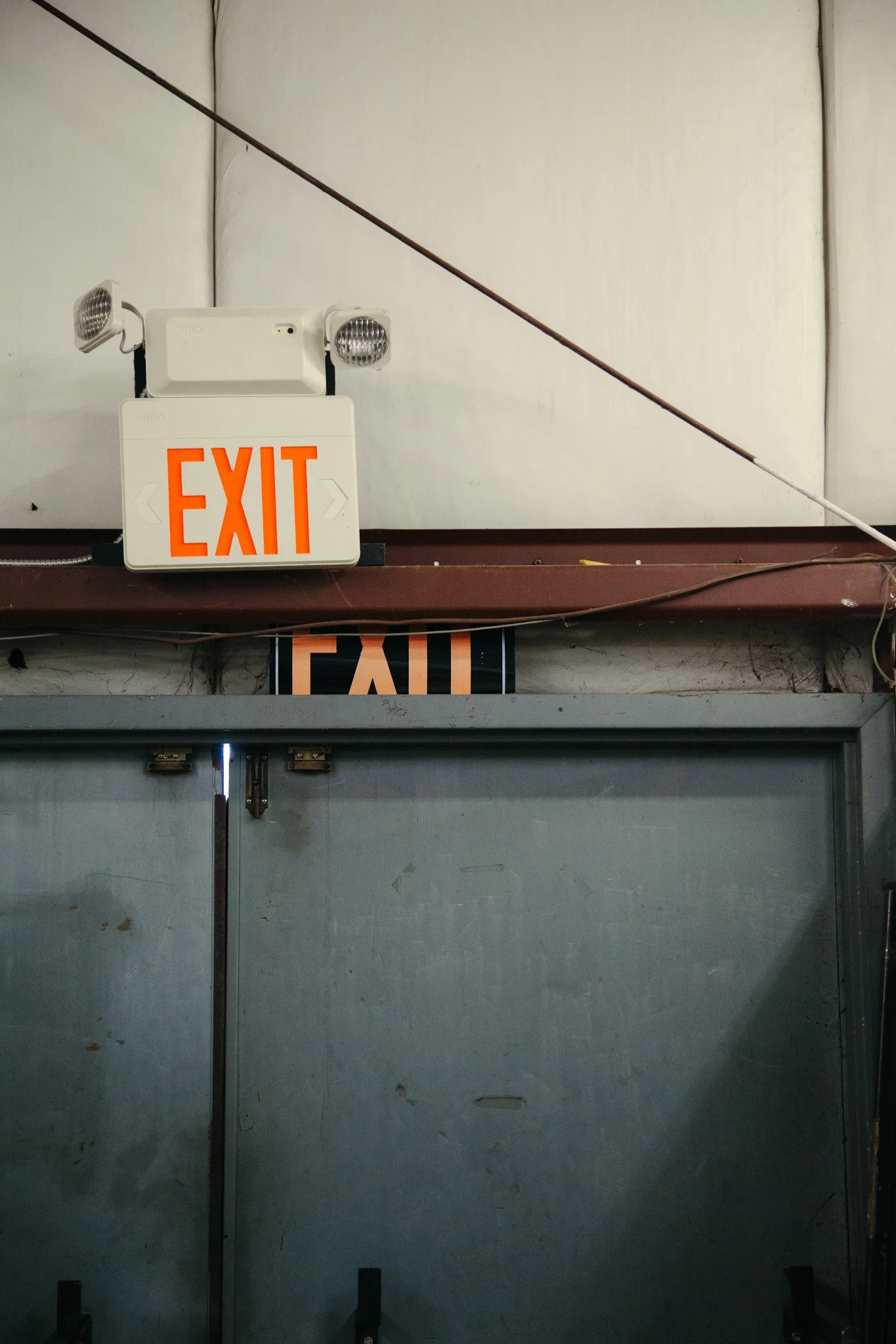 Industrial or commercial outdoor door with a large illuminated exit sign above it, and two security cameras mounted on the wall nearby.