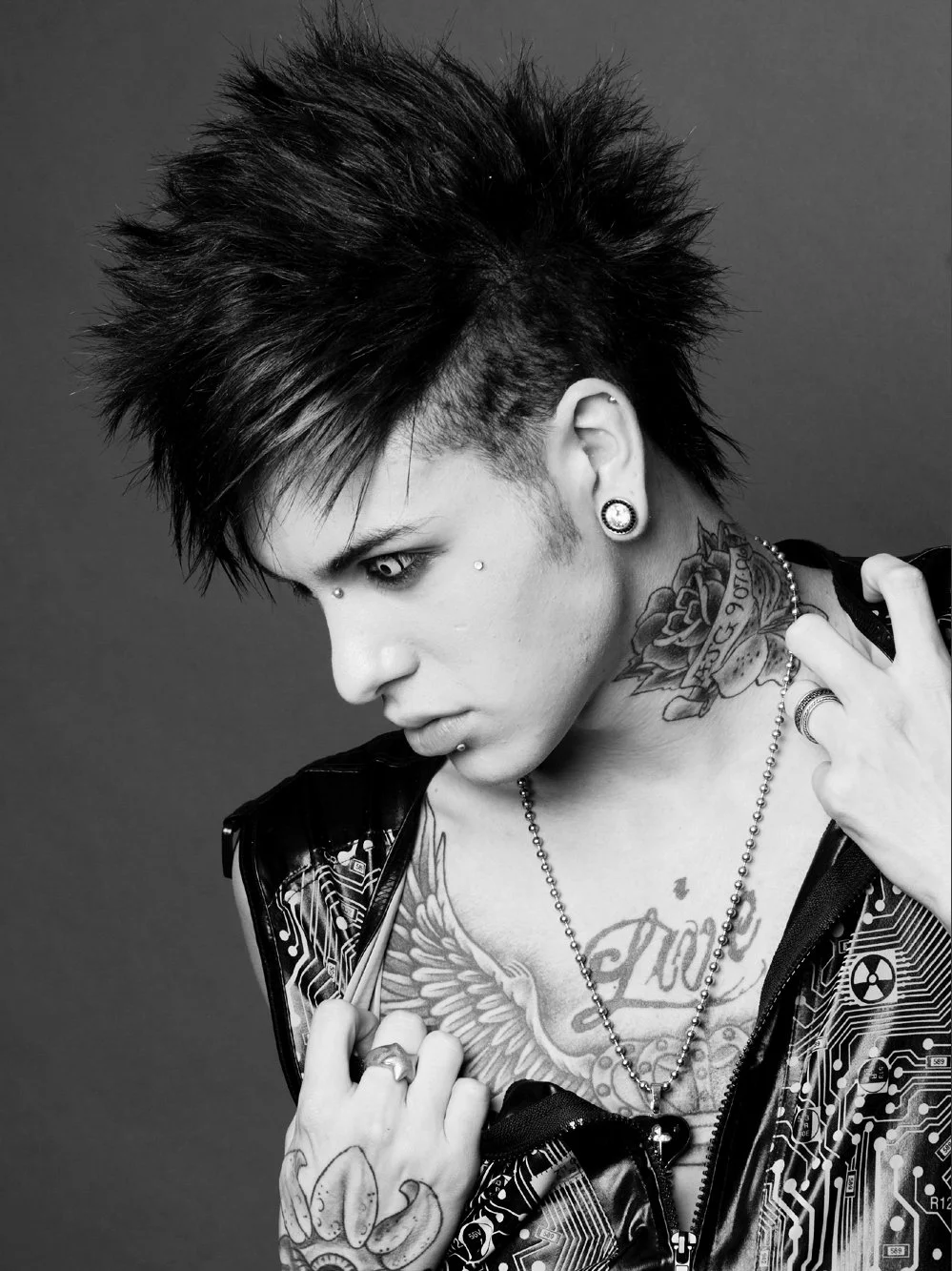 Black and white photo of a person with a mohawk hairstyle, tattoos on their chest and neck, piercings on their face and ears, wearing a jacket with circuit board patterns, and posing with their hand near their shoulder.