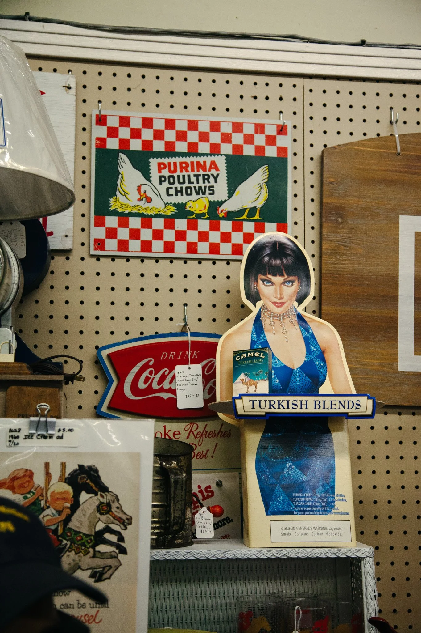 A vintage-style cardboard cutout of a woman wearing a blue dress with a necklace, promoting Turkish coffee blends, surrounded by other memorabilia and signs on a store display wall.