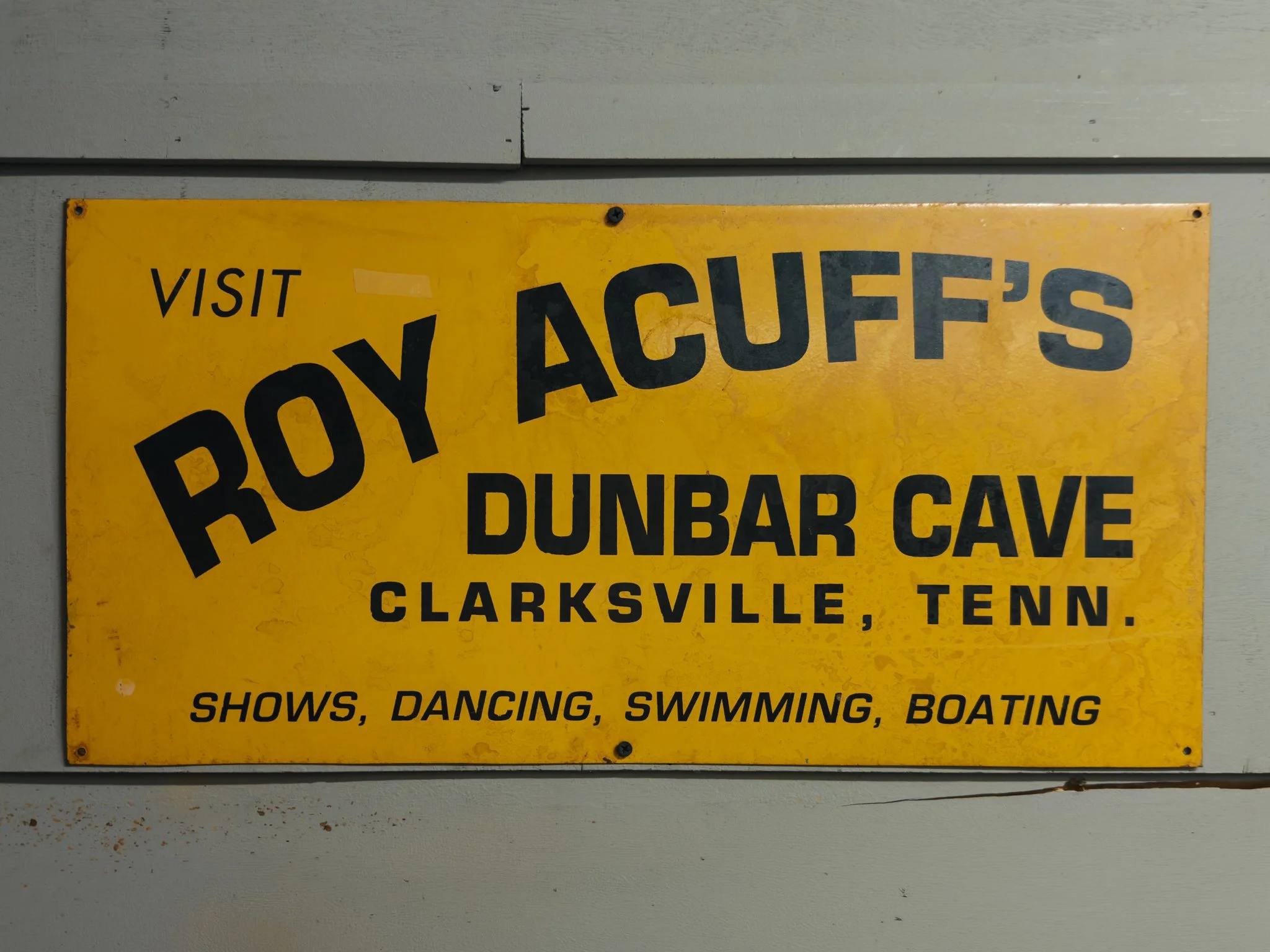 A yellow sign that reads 'Visit Bob Ack's, Boyd, Dunbar Cave, Clarksville, Tenn. Shows, dancing, swimming, boating' attached to a light gray wall.