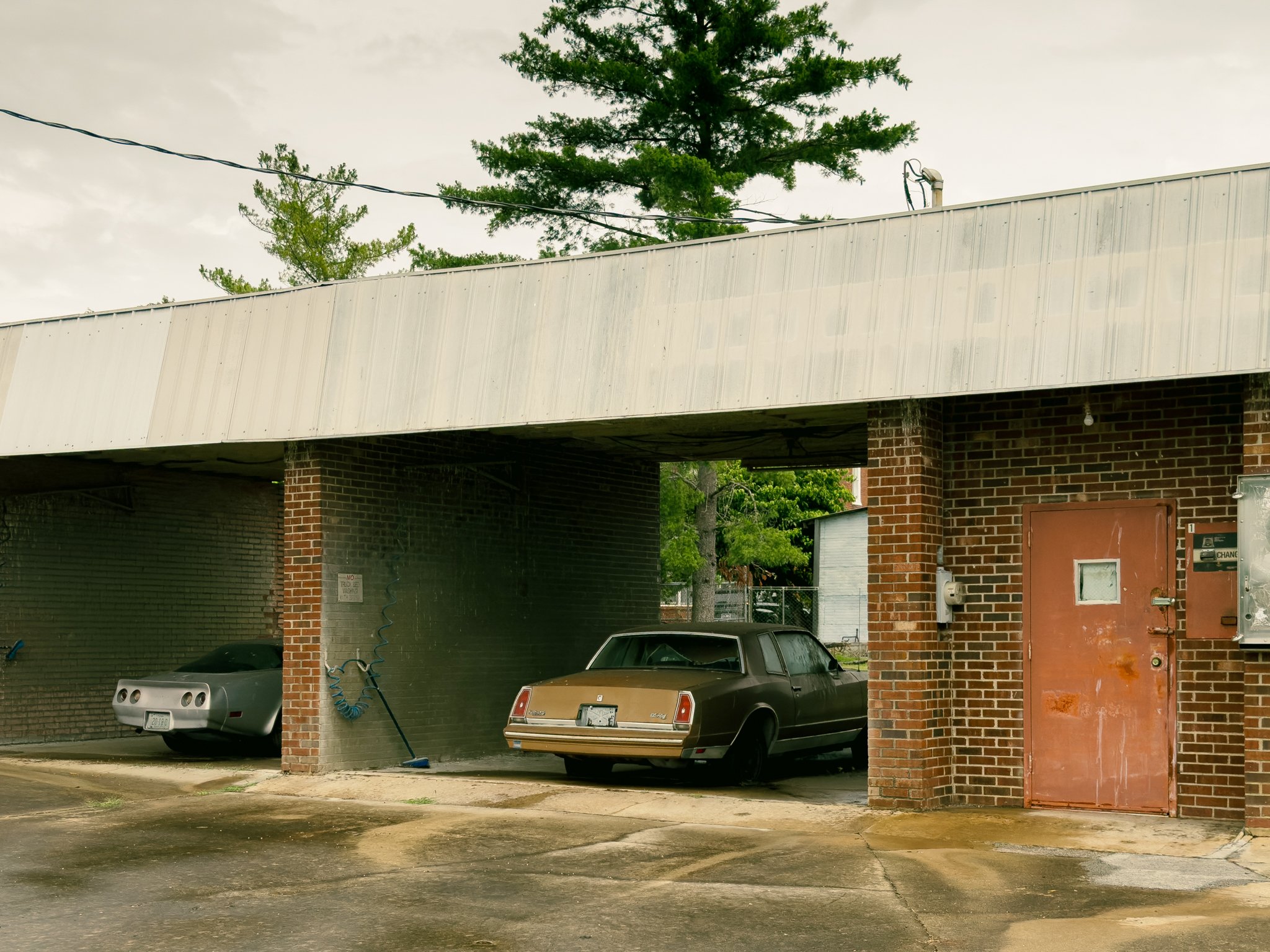 A parking area under a building with brick columns and a white roof. Two cars are parked underneath: a silver sports car on the left and a brown sedan on the right. There is a blue hose hanging from the ceiling, and the ground appears wet. In the bac