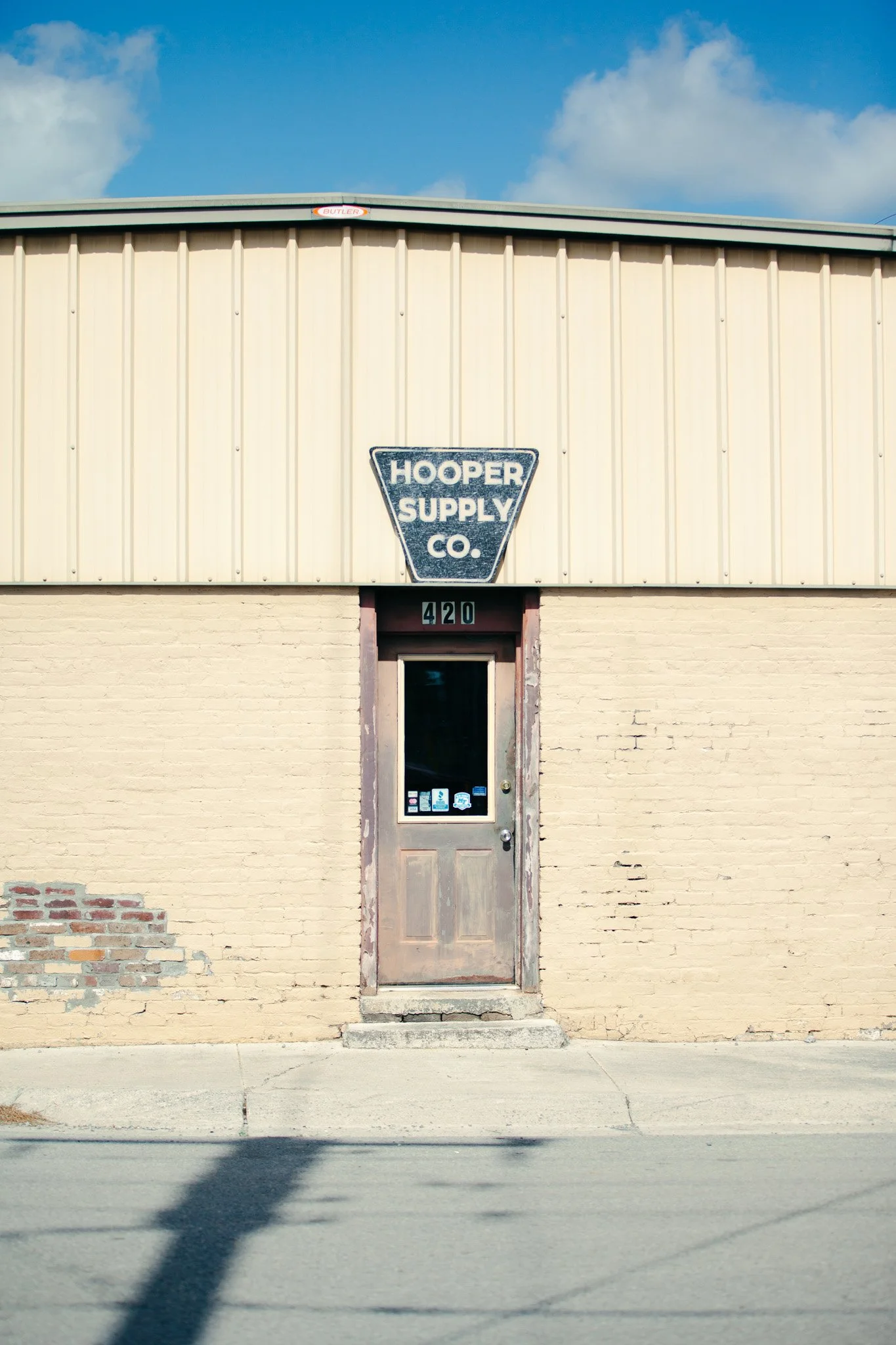 A building with a beige brick lower section and a tan upper section with vertical siding. There is a door with a small window and various stickers, and above it is a sign reading "Hooper Supply Co." The number 420 is above the door. The sky is blue w