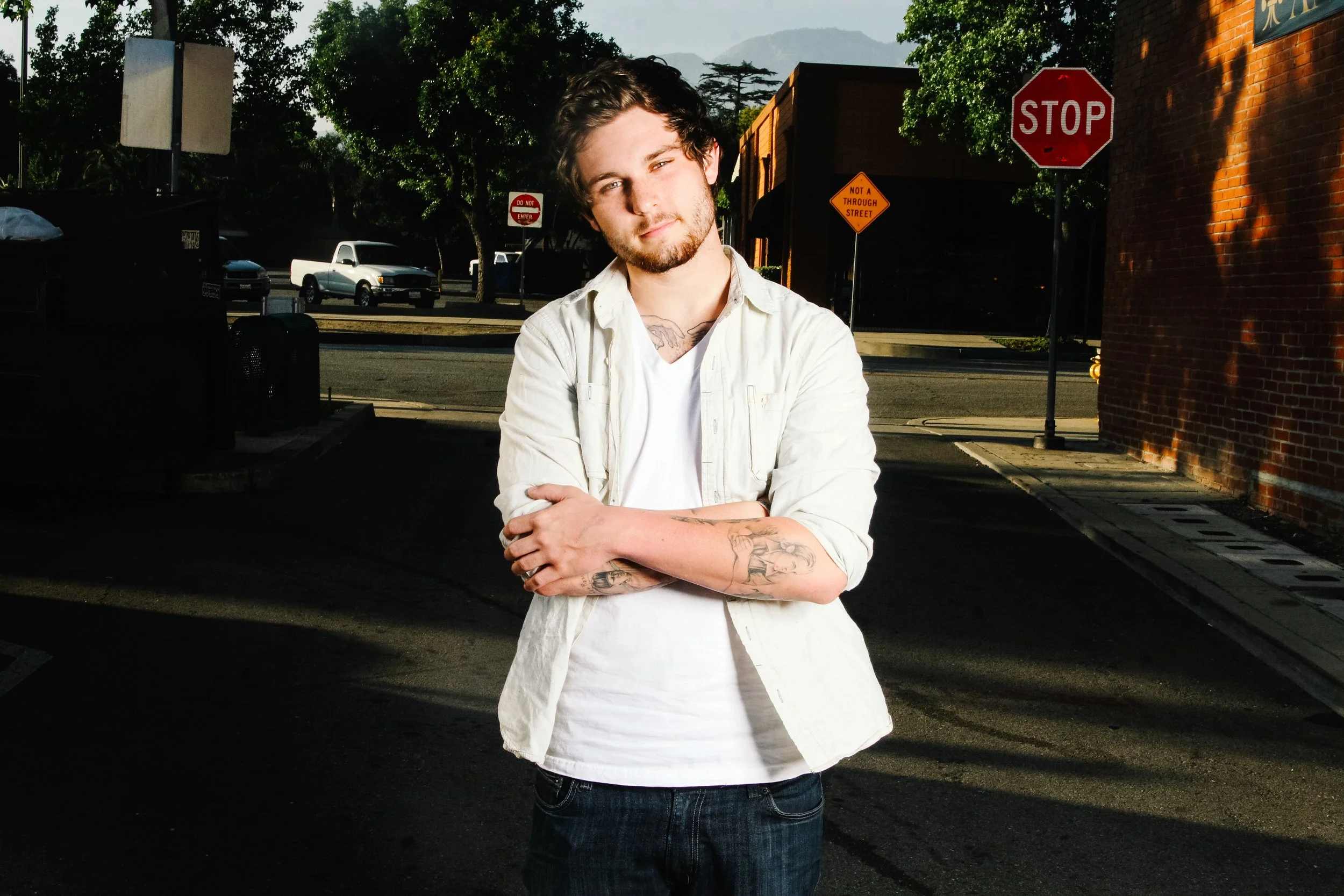 Young man with tattoos on his arms and chest, standing outdoors in front of a street scene, wearing a white shirt with rolled-up sleeves and dark jeans, with trees, parked cars, and stop signs in the background.