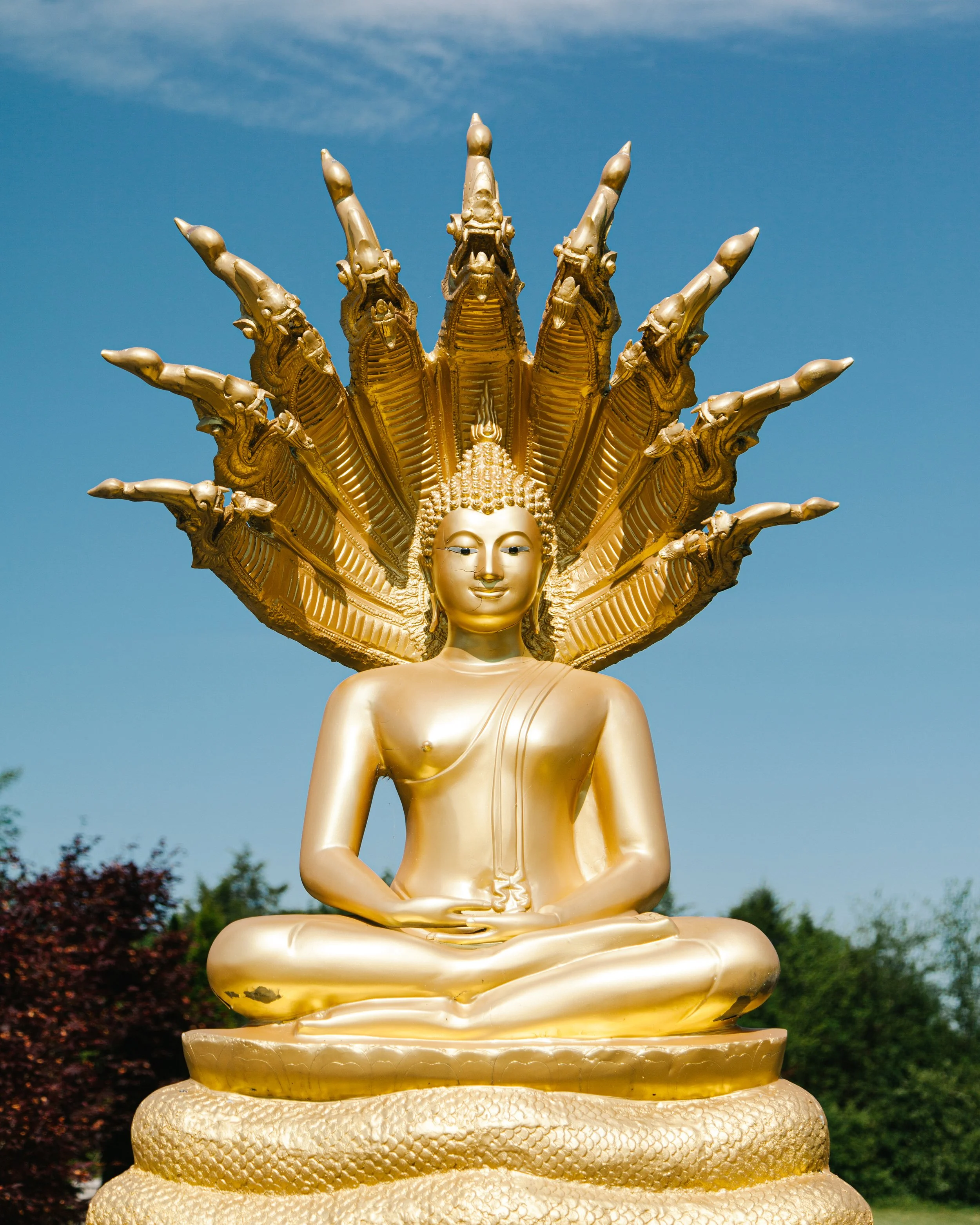 A golden Buddha statue with multiple snake heads forming a protective halo, set against a blue sky with some clouds.