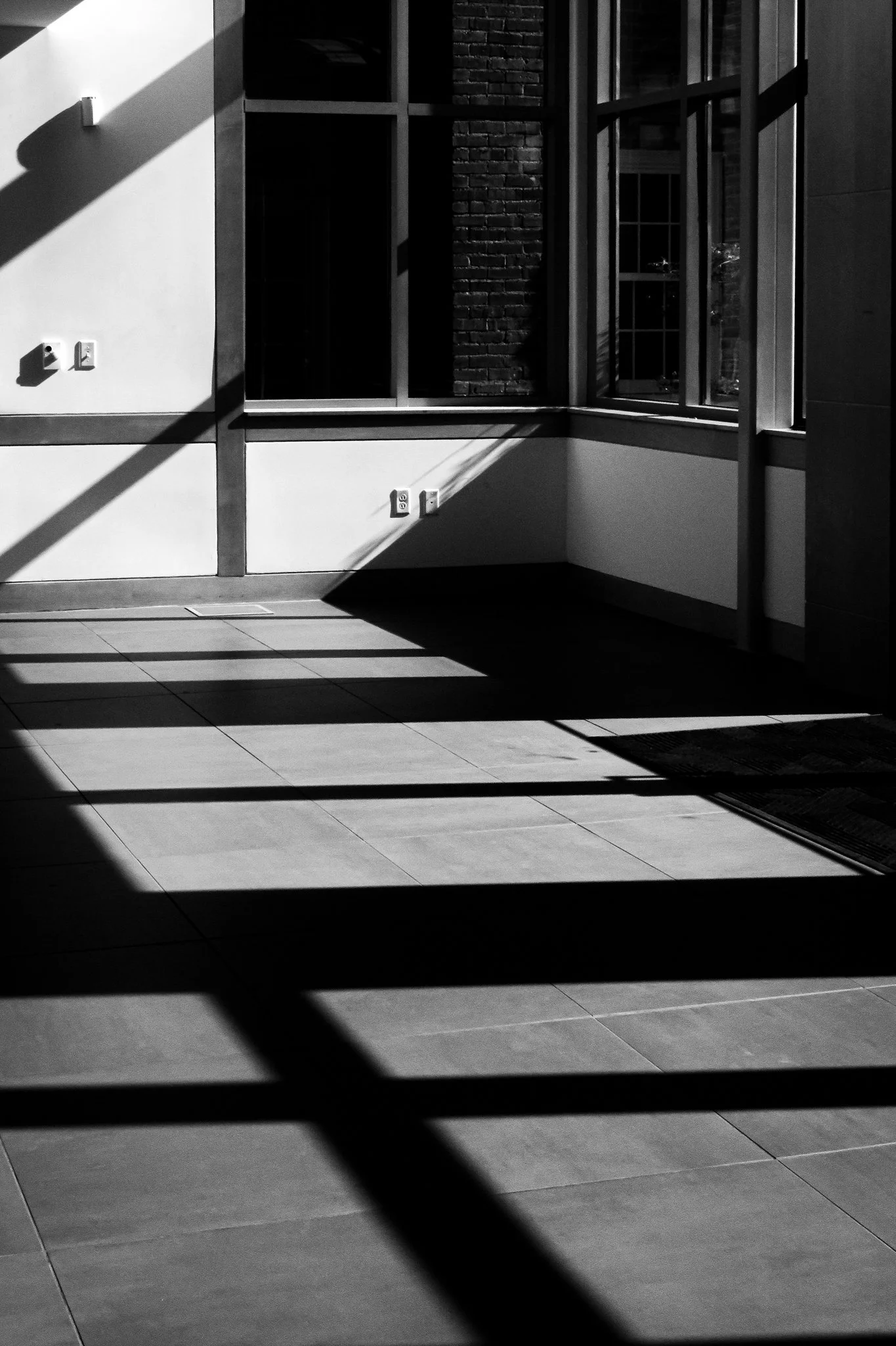 The image shows a modern interior space with sunlight streaming through large windows, casting shadows on the tiled floor. There are electrical outlets and wall fixtures visible on the white wall.