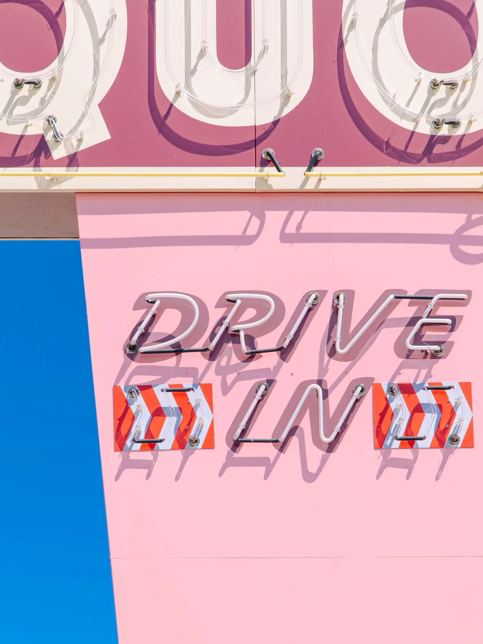 Close up of a pink wall with a colorful neon sign that reads 'DRIVE' in cursive, with decorative arrow elements and shadow effects.