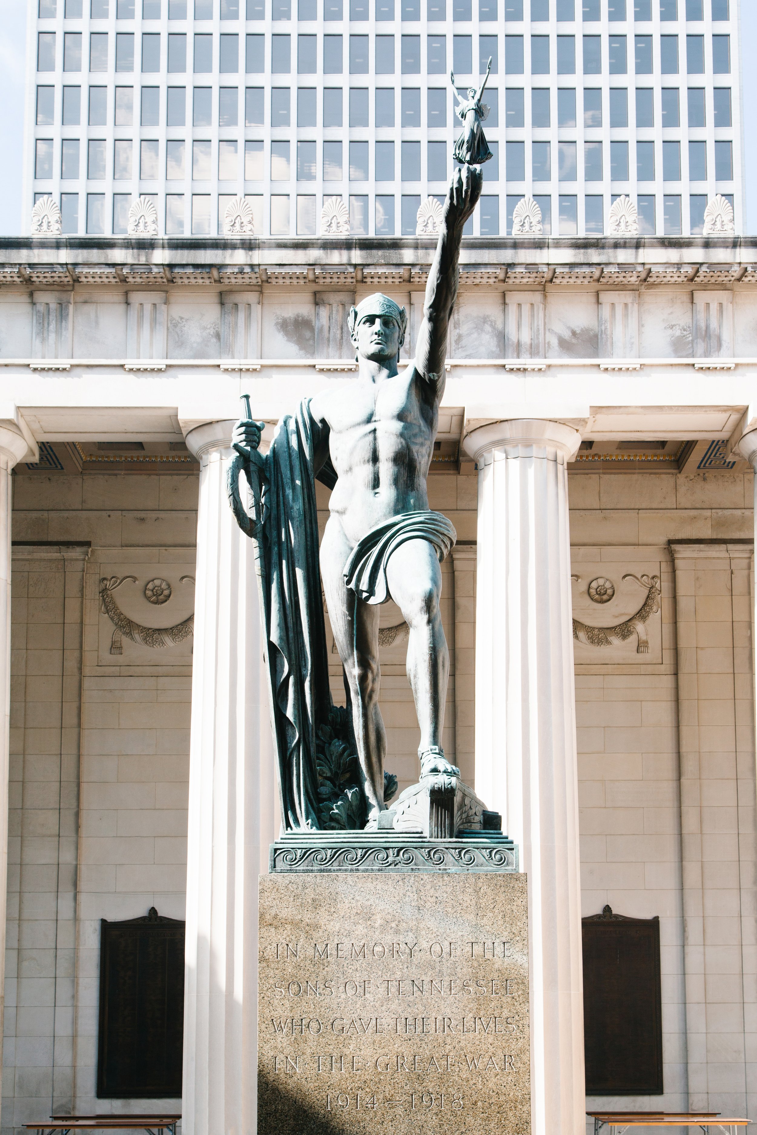 A statue of a soldier holding a sword and a shield, standing in front of a building with tall columns and a modern glass facade. The base of the statue has an inscription in memory of Sons of Tennessee who gave their lives in the Great War (World War