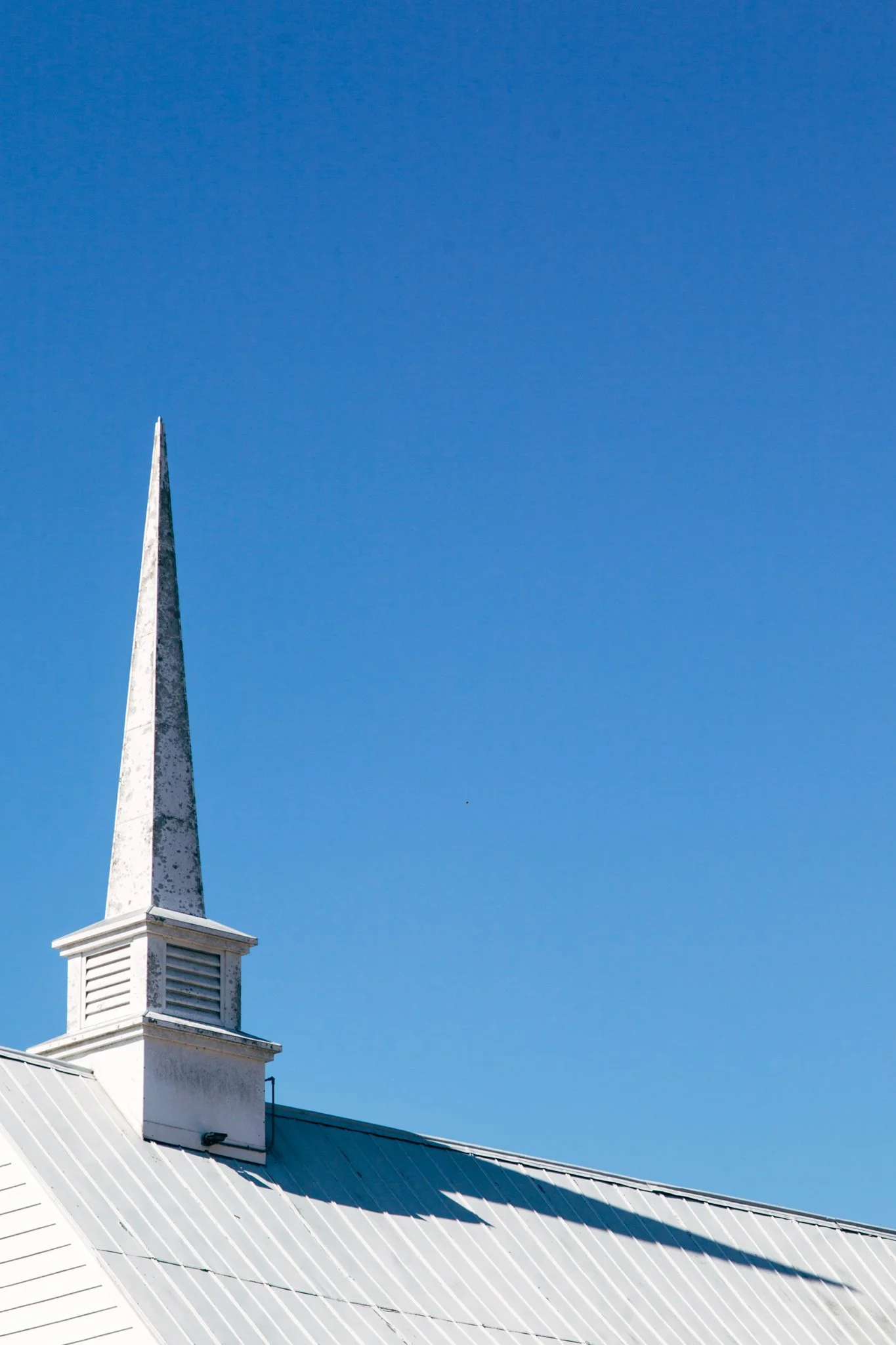 White church steeple with a pointed spire and a small box-shaped top, set against a clear blue sky, attached to a white building with a metal roof.