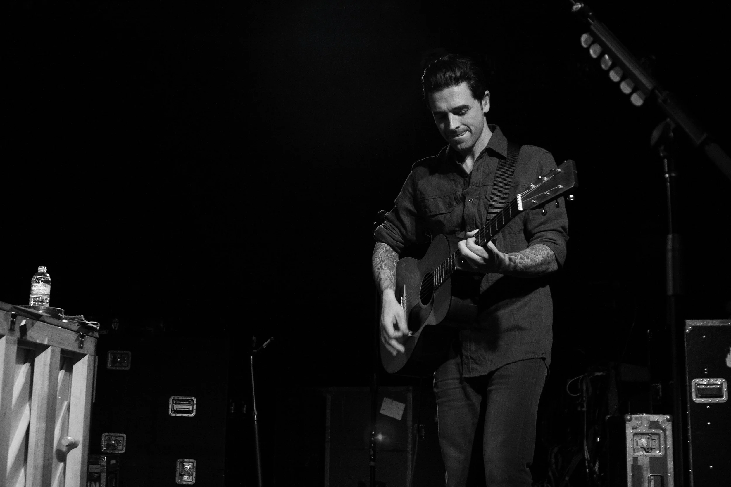 A man playing an acoustic guitar on stage, with a dark background and a music stand to his right.