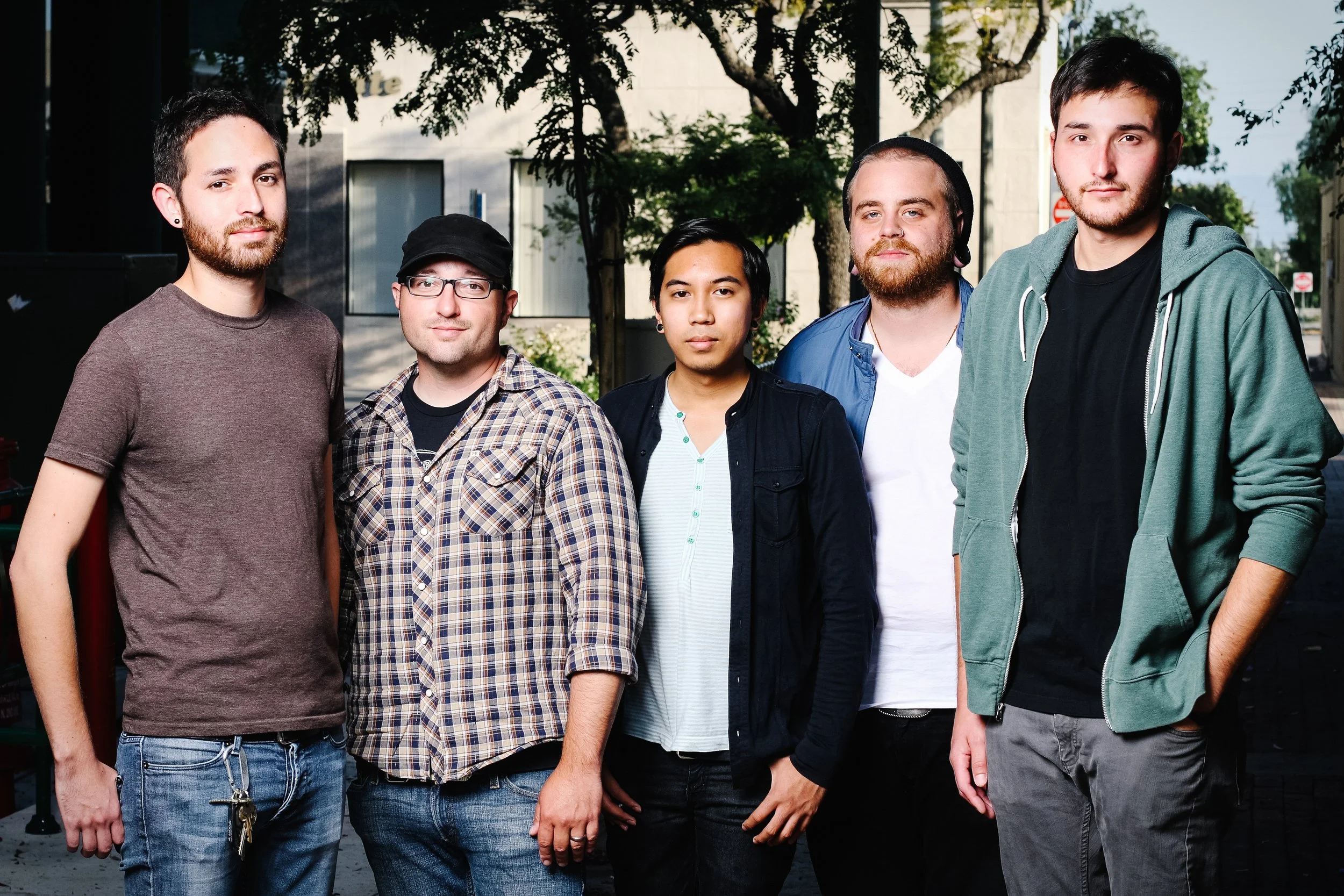Group of six young men standing outdoors in front of trees and buildings, dressed casually.