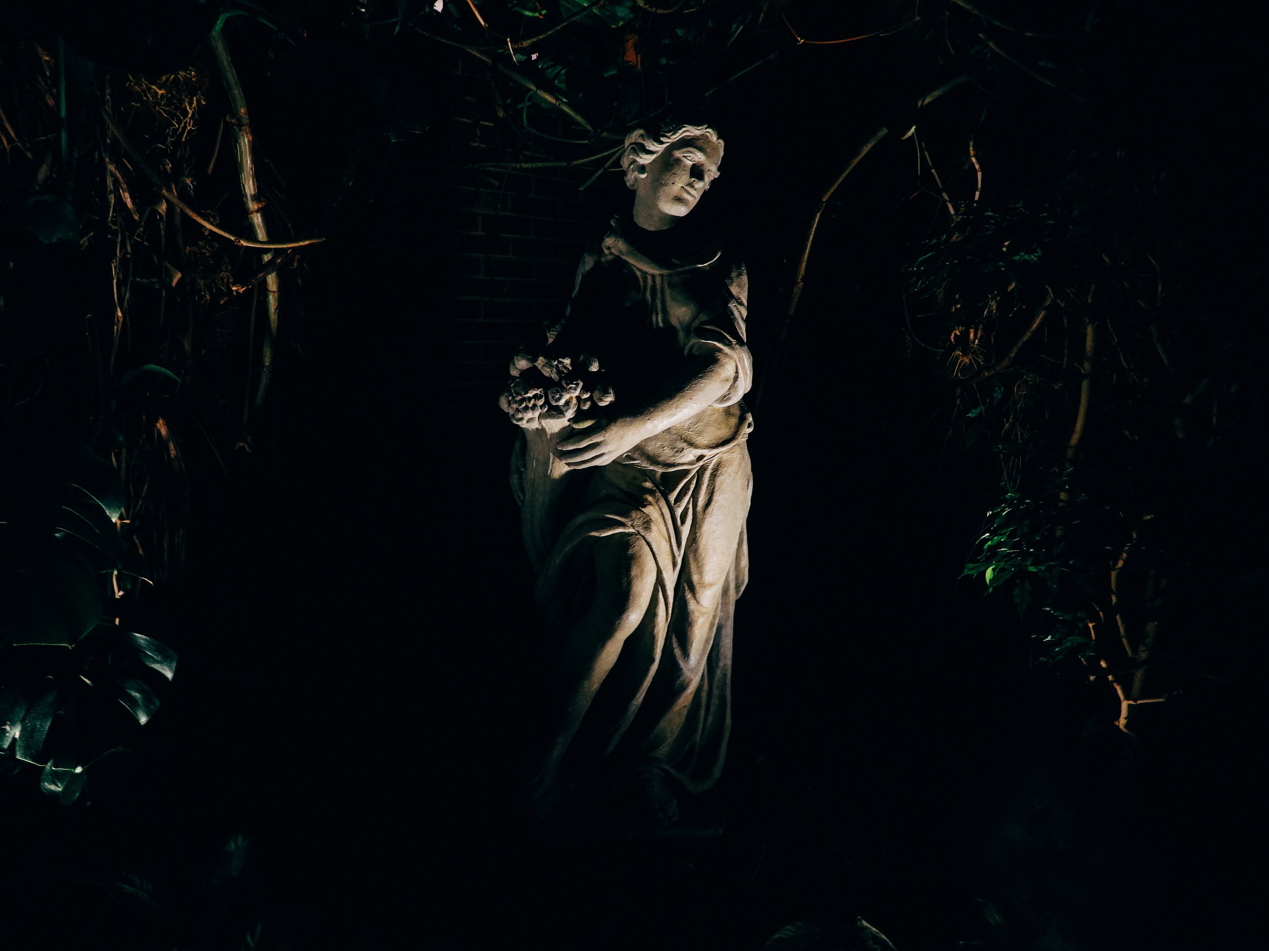 A stone sculpture of a woman with flowing hair, standing outdoors at night surrounded by dense vegetation.