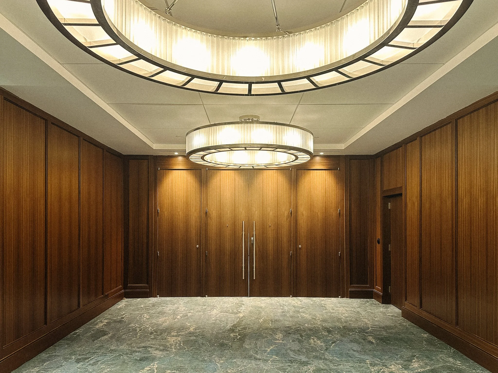 This image shows an elegant, modern room with dark wood paneled walls, gray marble flooring, and two large, circular chandeliers hanging from the ceiling, illuminating the space.
