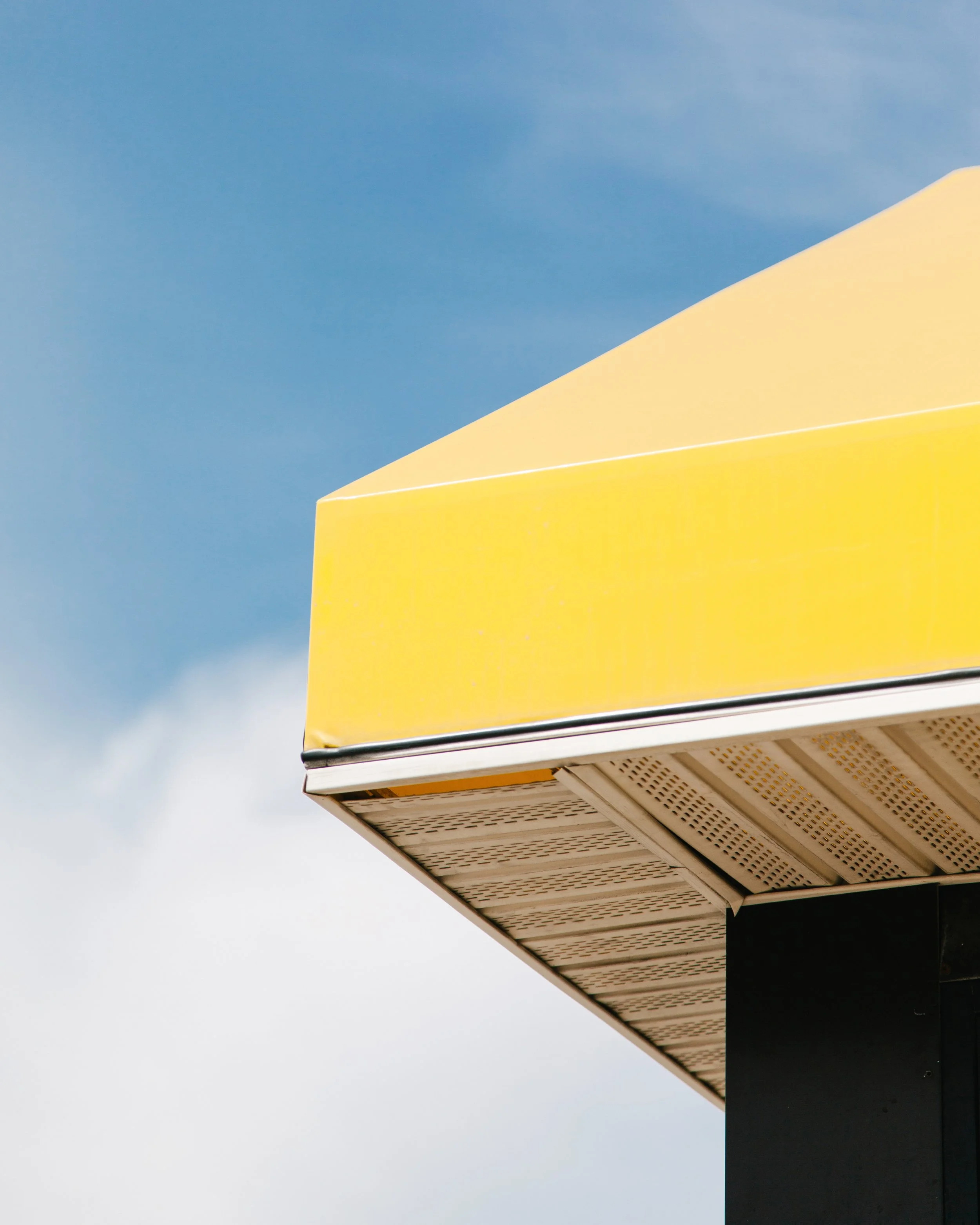 Close-up of a building corner with a bright yellow section, beige details, and a black section below against a partly cloudy sky.