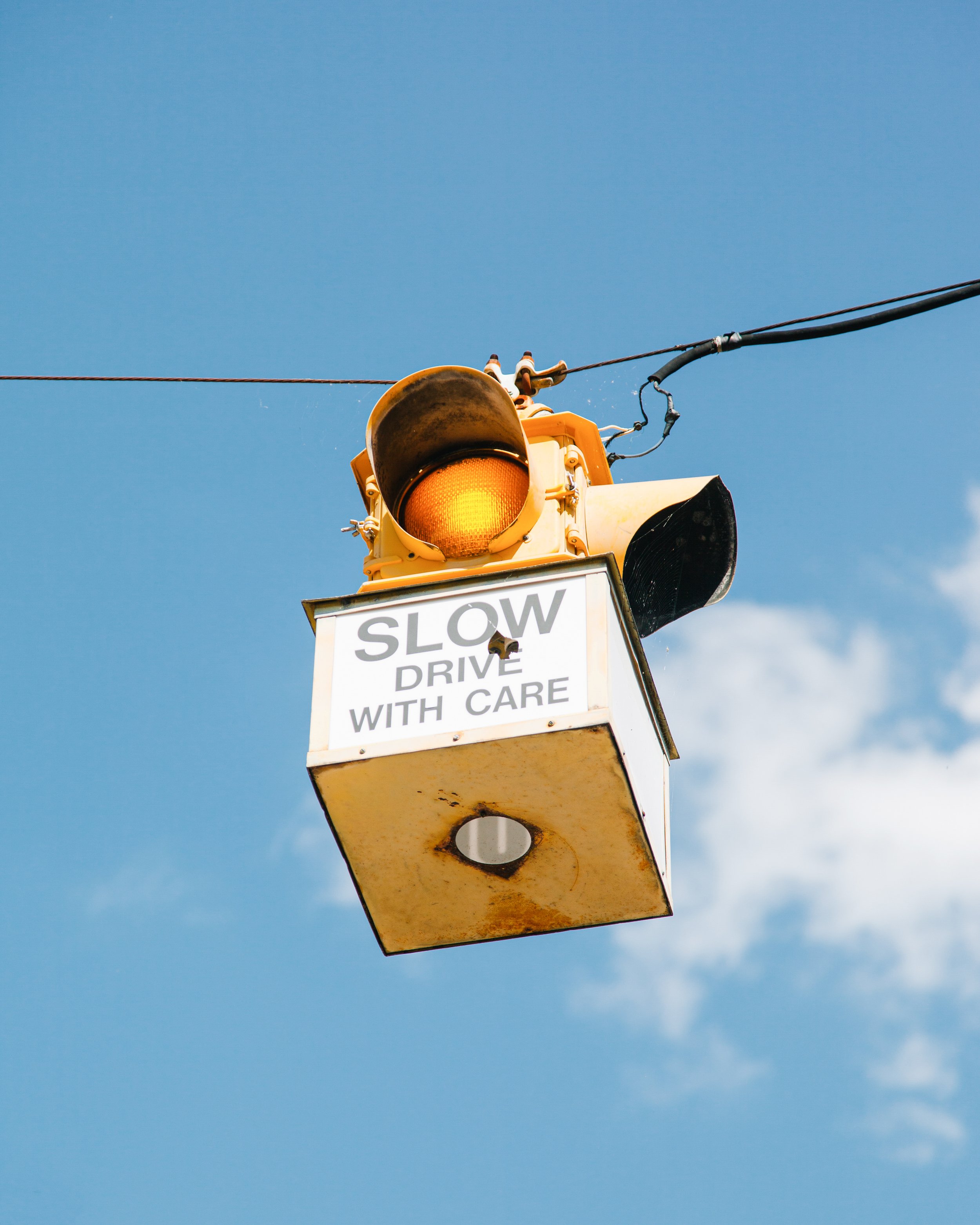 Yellow traffic light mounted on a pole against a blue sky, with a partly visible sign that reads 'SLOW'.