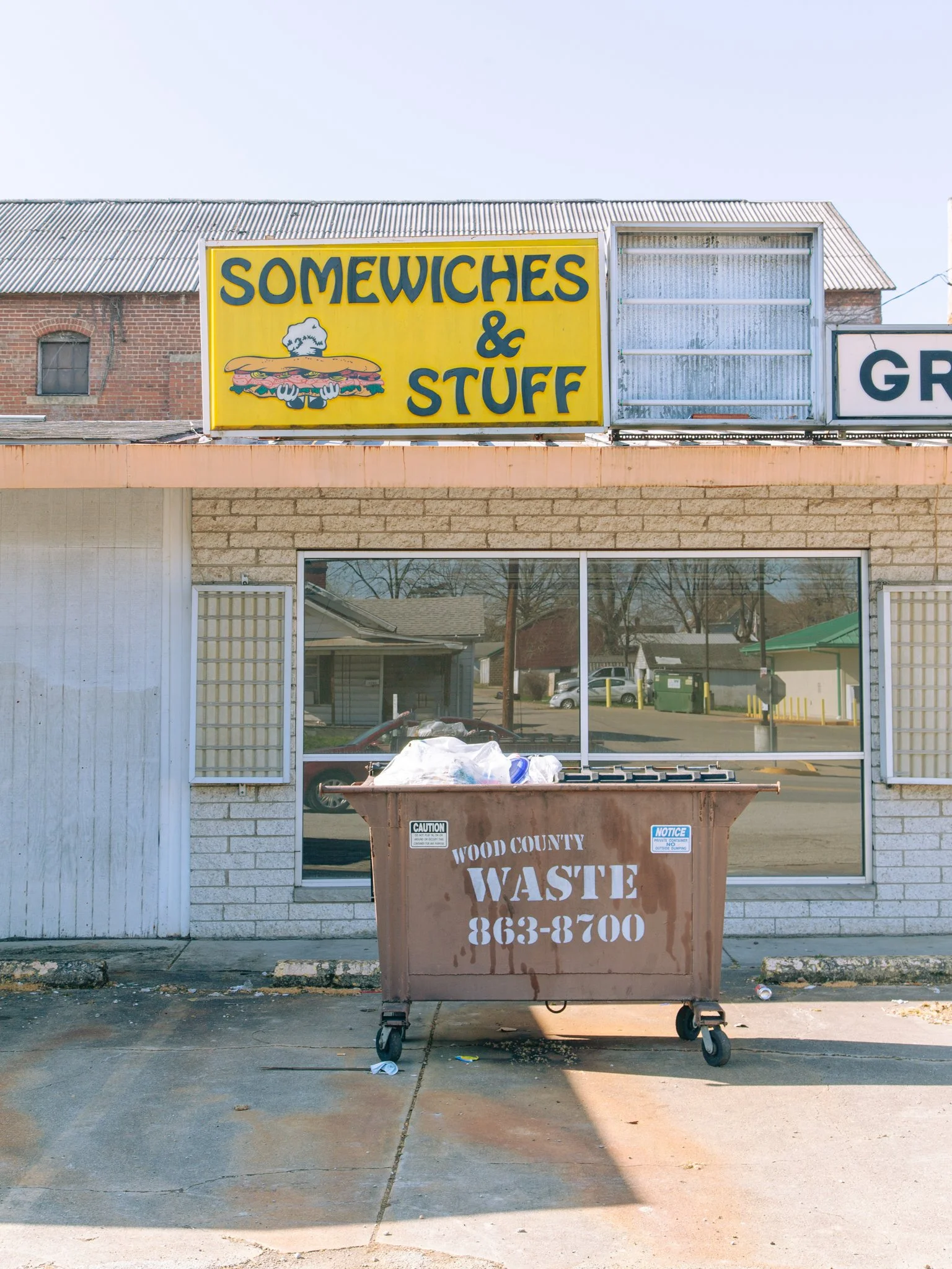 A yellow sign with blue letters reads 'Somewiches & Stuff' with a cartoon of a sandwich with smoke coming out of it. Below, a brown dumpster labeled 'Wood County Waste 863-8700' is overflowing with trash outside a building with large windows and a br