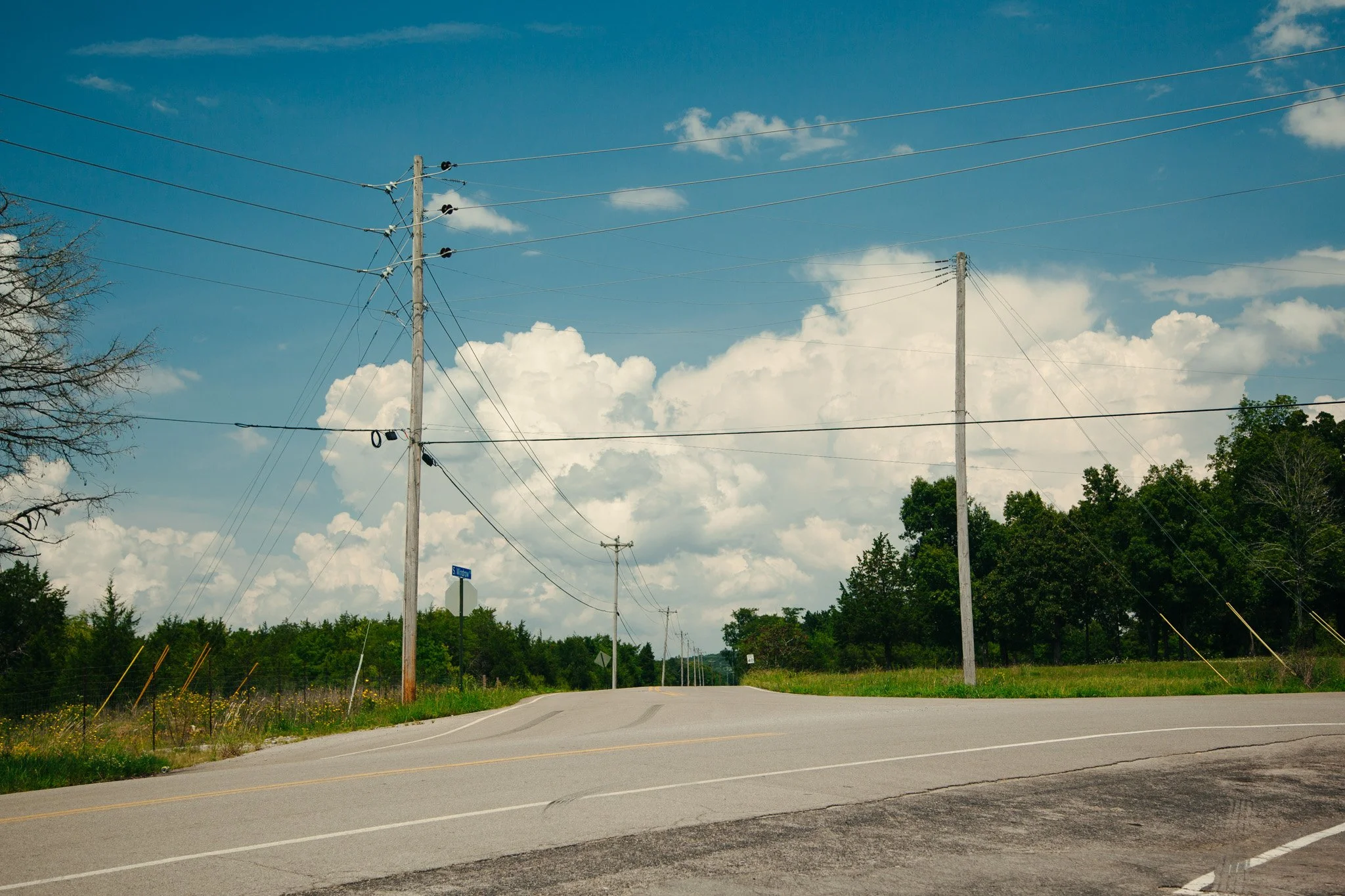 A rural road with utility poles and power lines, with trees and a partly cloudy sky in the background.