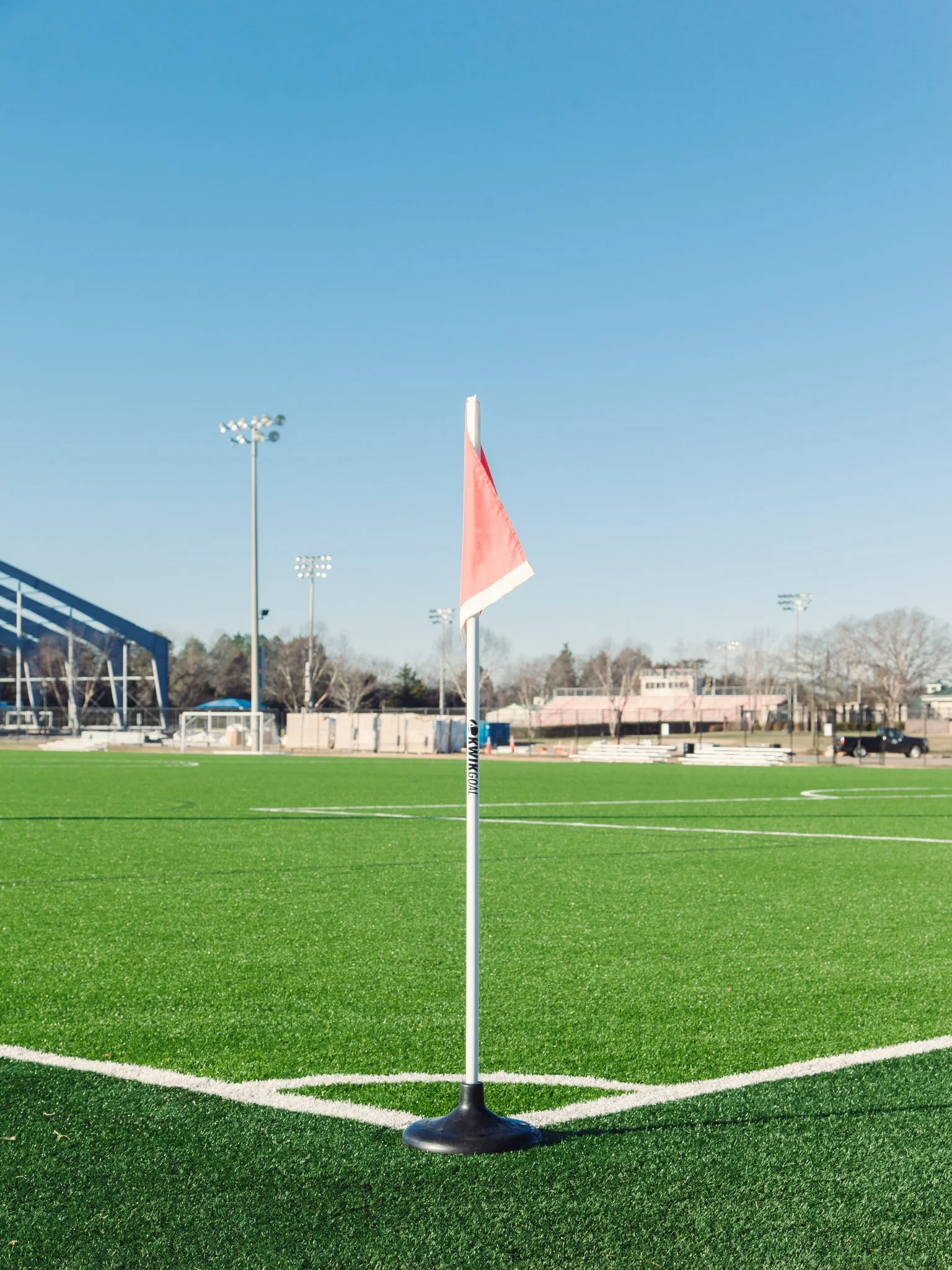An empty sports field with bright green artificial turf, a red and white flag on a pole in the center, and a clear blue sky overhead.