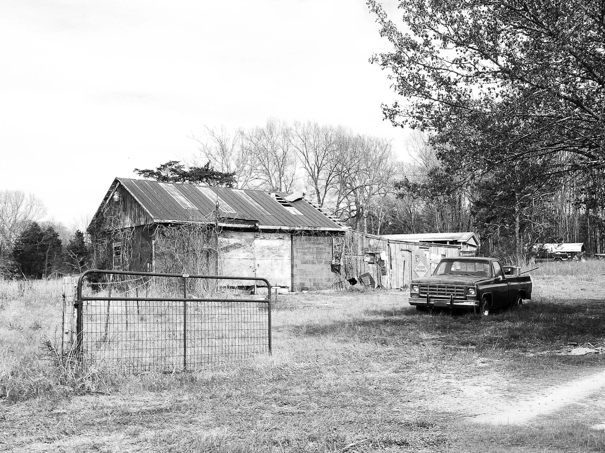 Black and white photo of a rustic barn with a corrugated metal roof, surrounded by leafless and evergreen trees, with a parked pickup truck in the foreground and a sign that reads 'Hidden Entrance'.