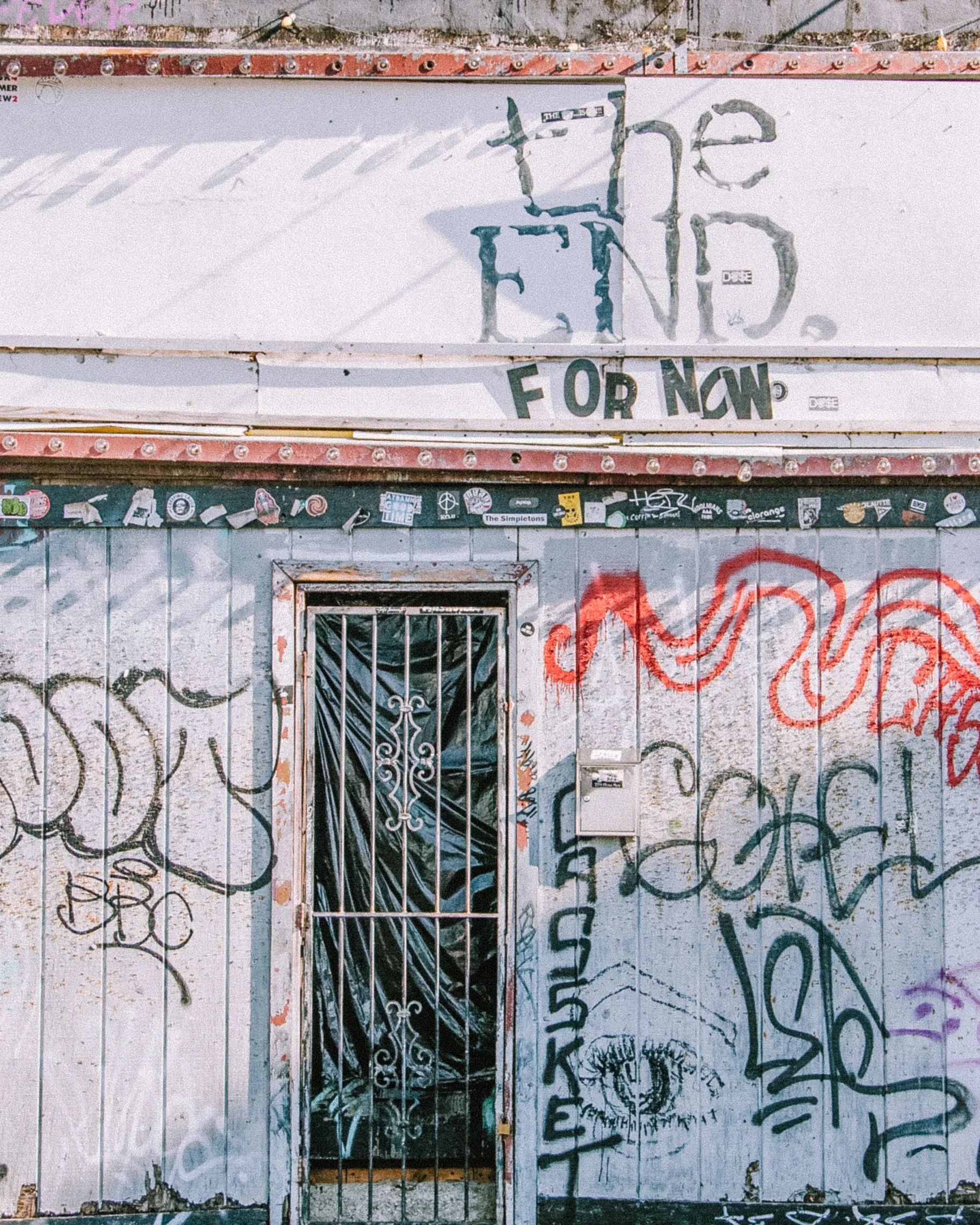 A metal gate with black paint peeling, set into a wall covered in graffiti. Above the gate, there is a sign with partially blocked text that reads 'the end for now' in painted and sticker letters. The wall has various tags, scribbles, and stickers, w