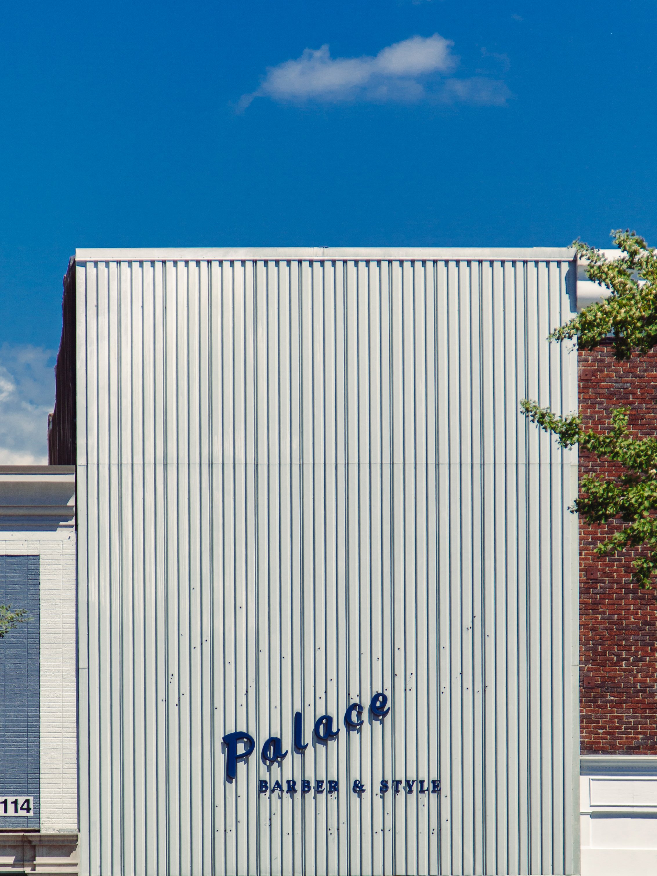 The exterior of a building with a white corrugated facade, part of a brick wall, and a blue sign that reads 'Palace Barber & Style'.