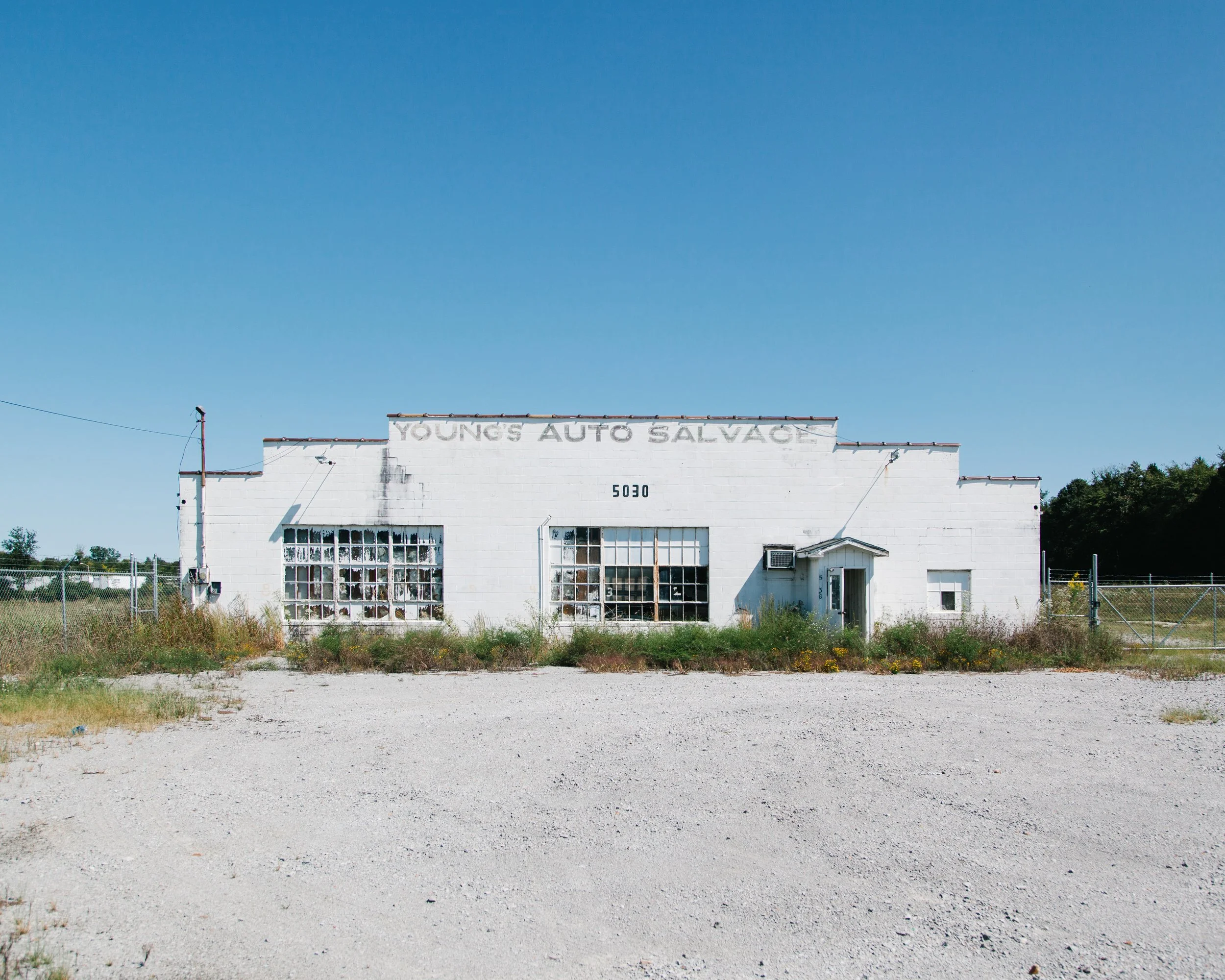 A simple white building with a sign that reads 'YOUNG'S AUTO SALVAGE' on a clear day with blue sky. The building has a chain-link fence in front and some overgrown grass around it.