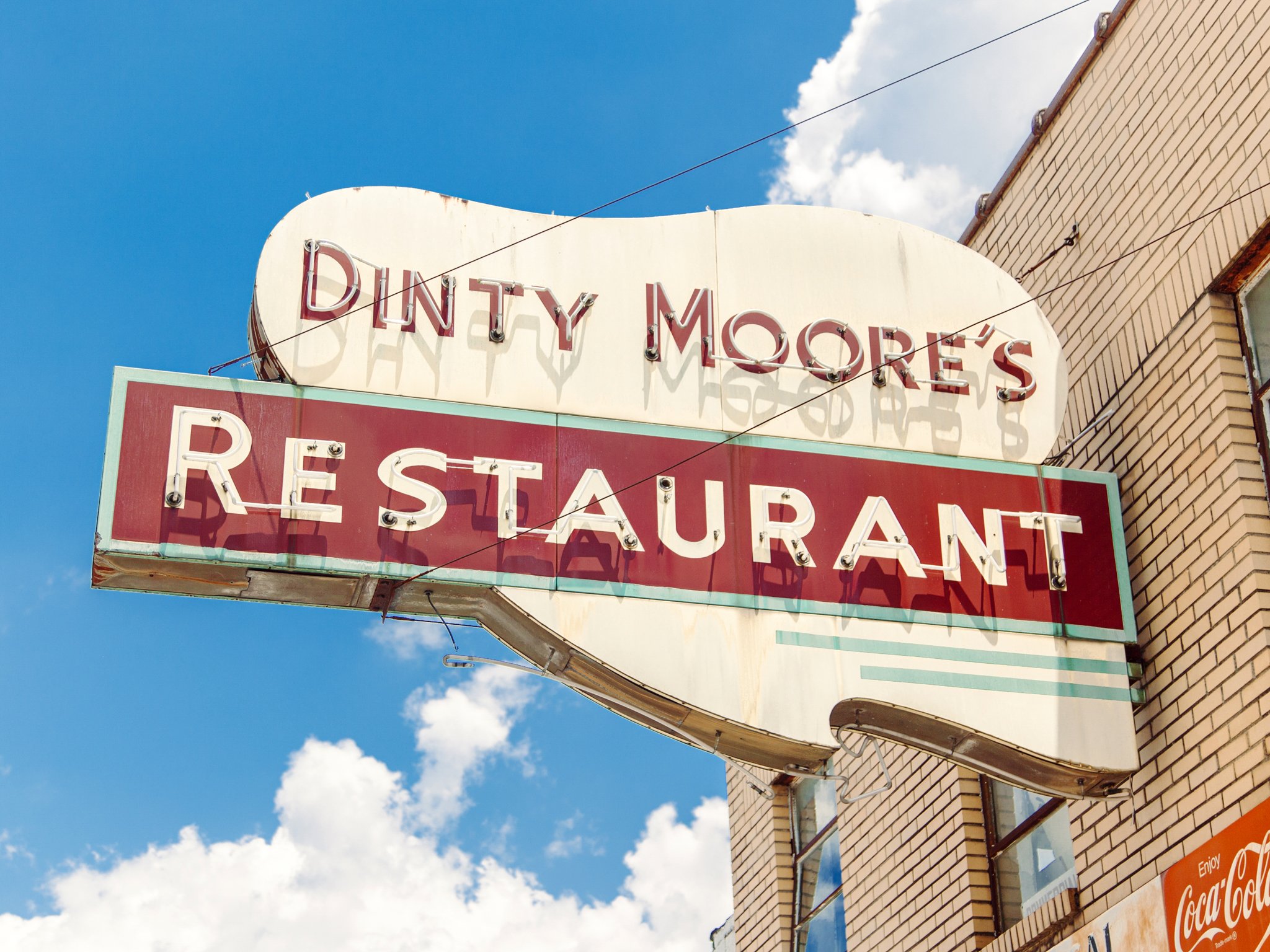 A vintage neon sign reading 'Dinty Moore's Restaurant' mounted on the side of a beige brick building against a blue sky with some white clouds.