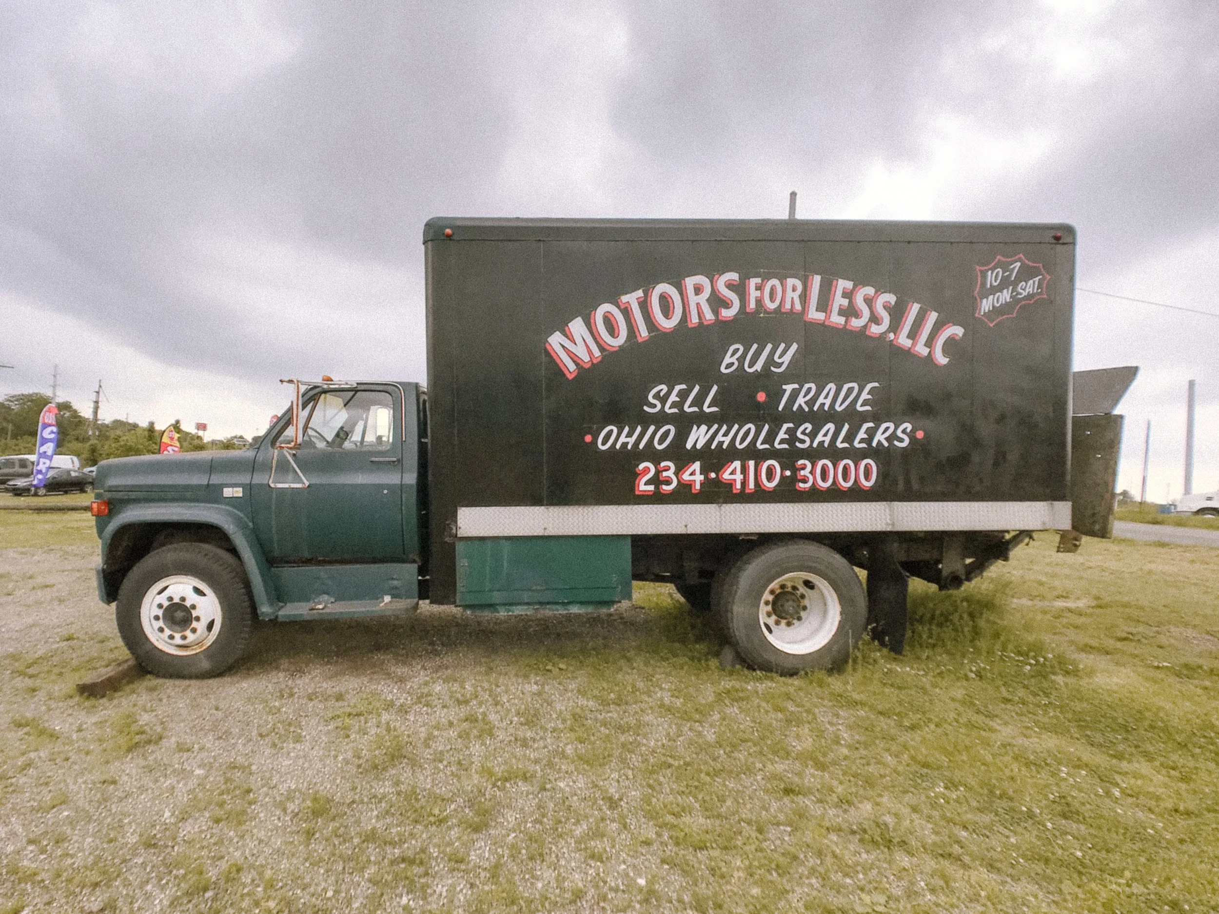A black truck with a large sign on its side advertising motors for less, with services like buy, sell, trade, and wholesale. The sign includes a phone number and business hours. The background shows cloudy skies and a parking lot with other vehicles 
