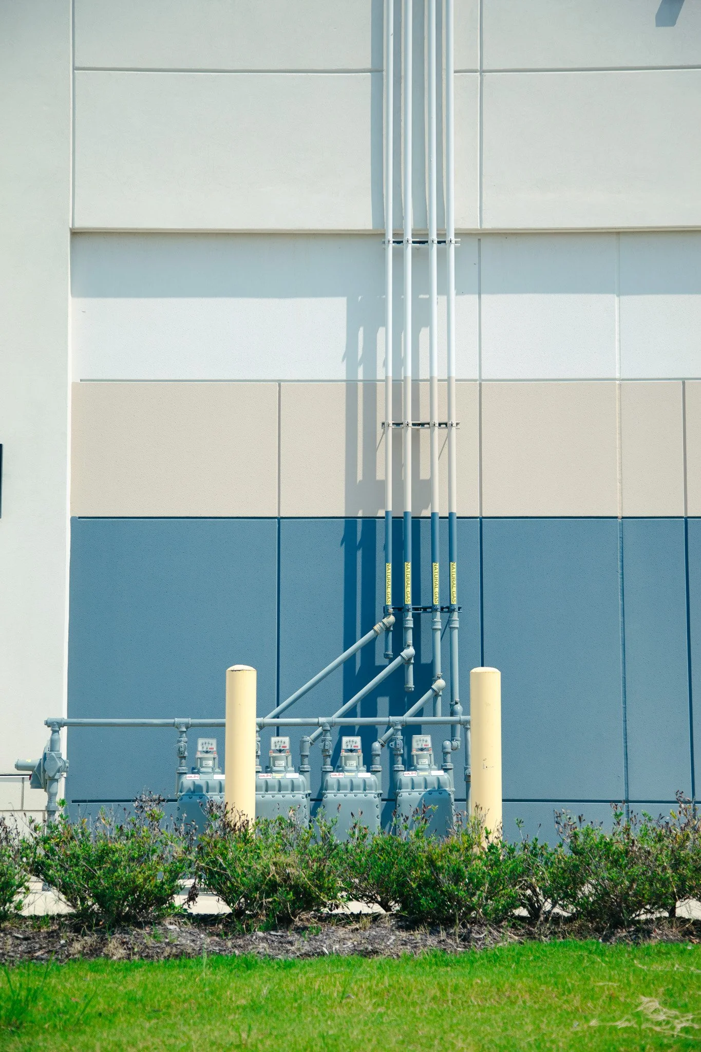 Utility building wall with pipes, meters, yellow bollards, and a row of bushes in front at Tanger Outlets in Antioch, TN
