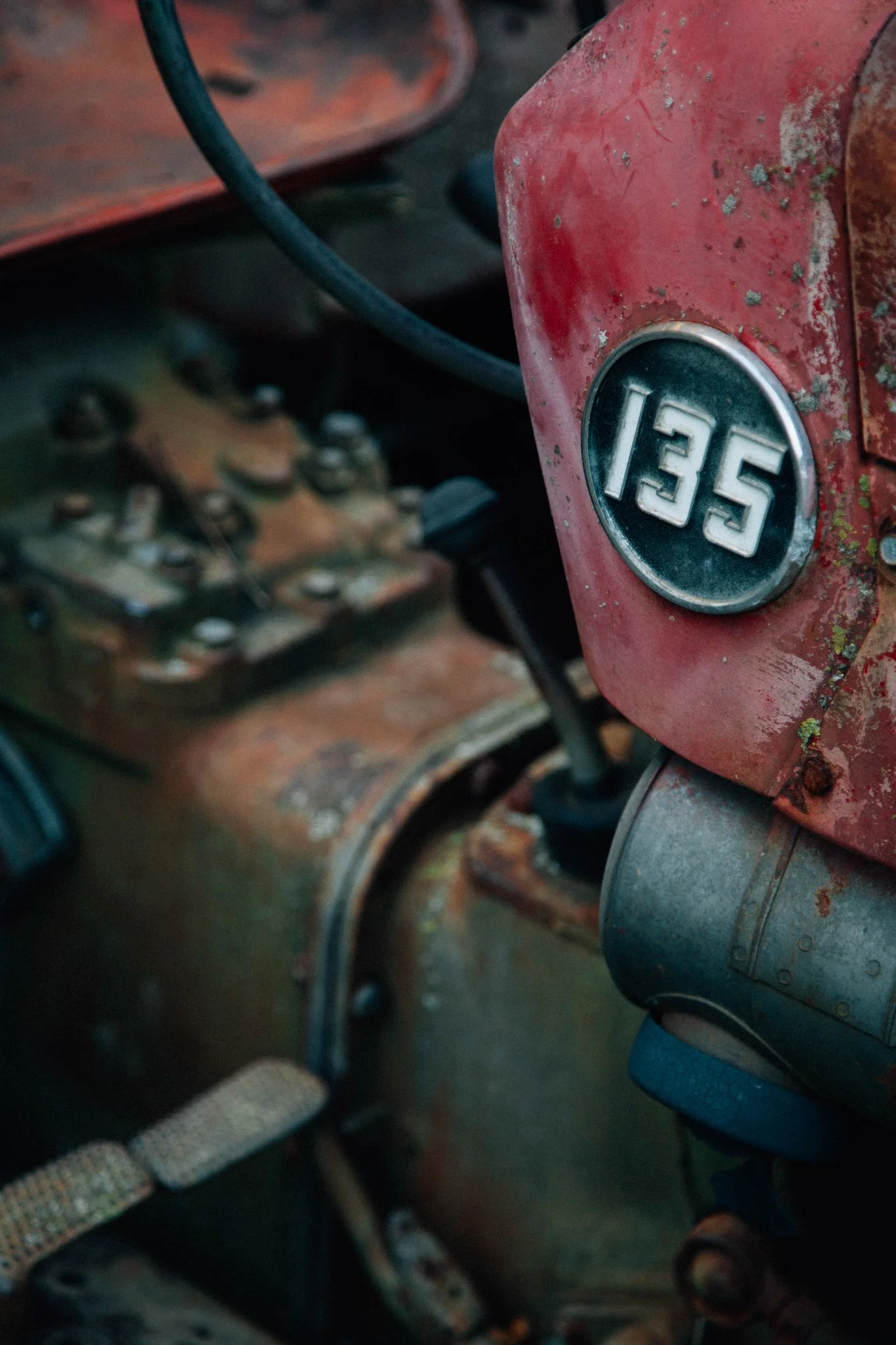Close-up of a vintage red tractor with the number '135' on a badge, showing signs of rust and wear, with parts of the machinery and a person’s leg and foot in the frame.