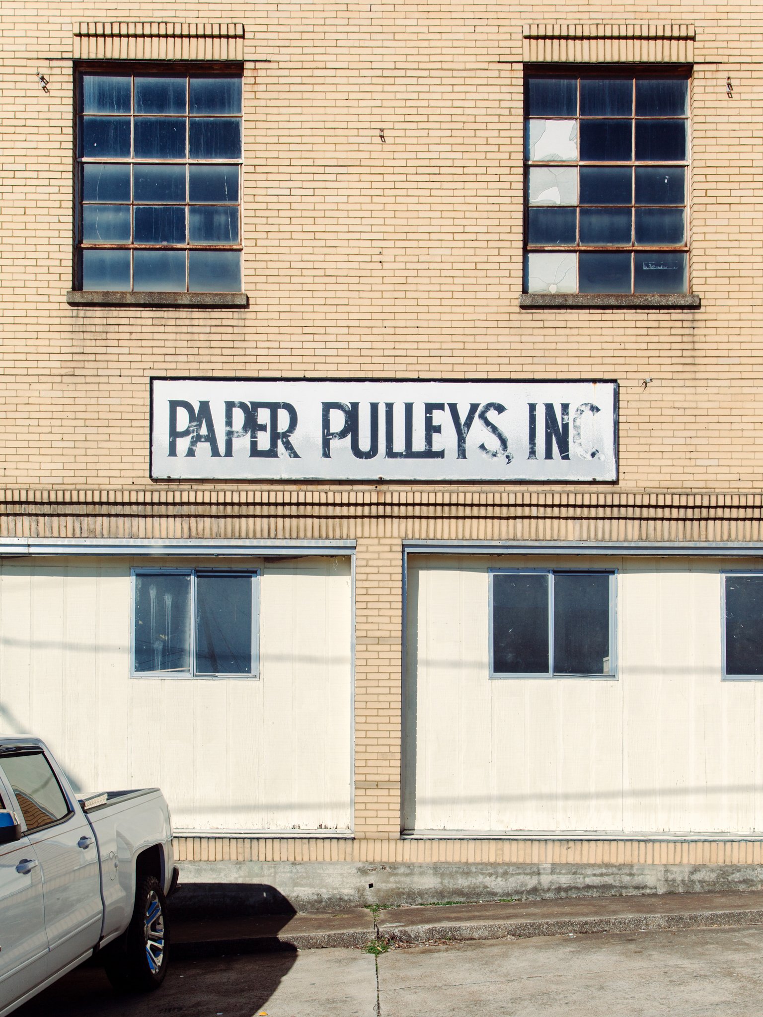 The image shows the exterior facade of a brick building with two large windows and an old, worn sign that reads "PAPER PULPYS INC" in faded black letters. There are some cars parked at the bottom of the image, and the setting appears to be in a parki
