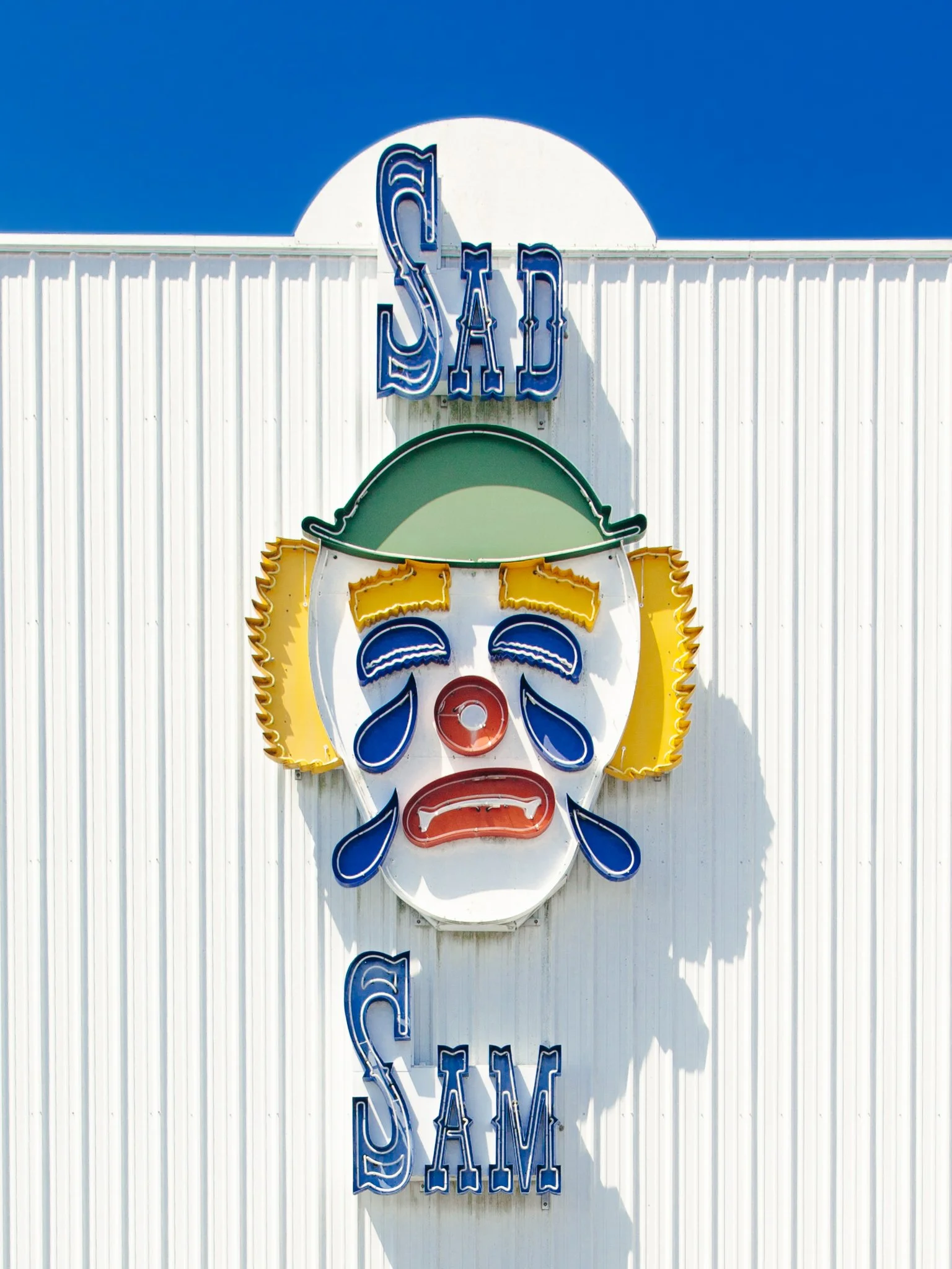 A neon sign of a clown face with the words 'Saloon' above it, mounted on a white corrugated metal wall against a blue sky.
