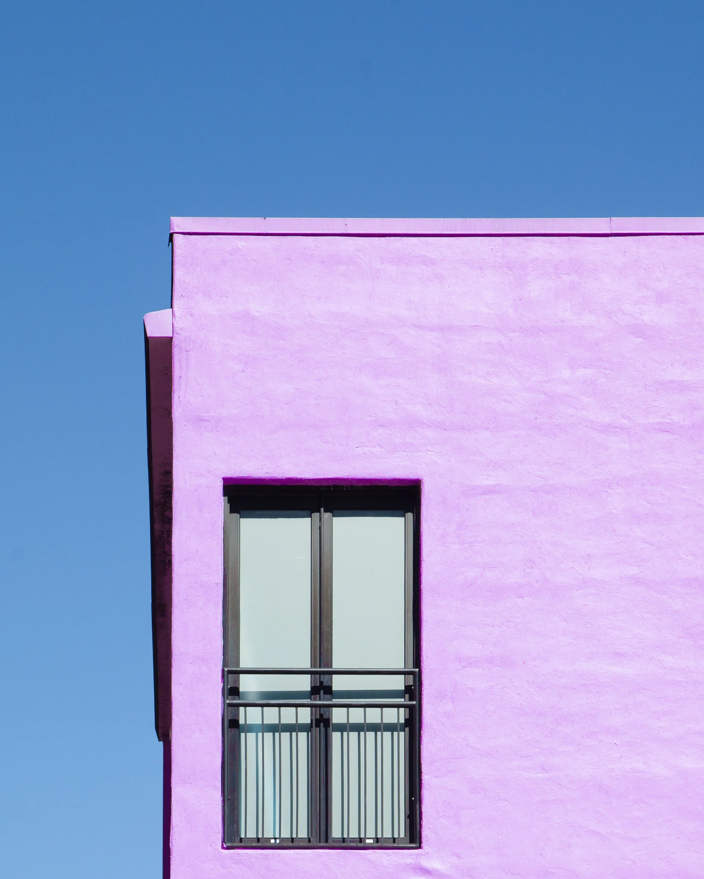 A pink building with a single window and a balcony with metal rails, set against a clear blue sky.