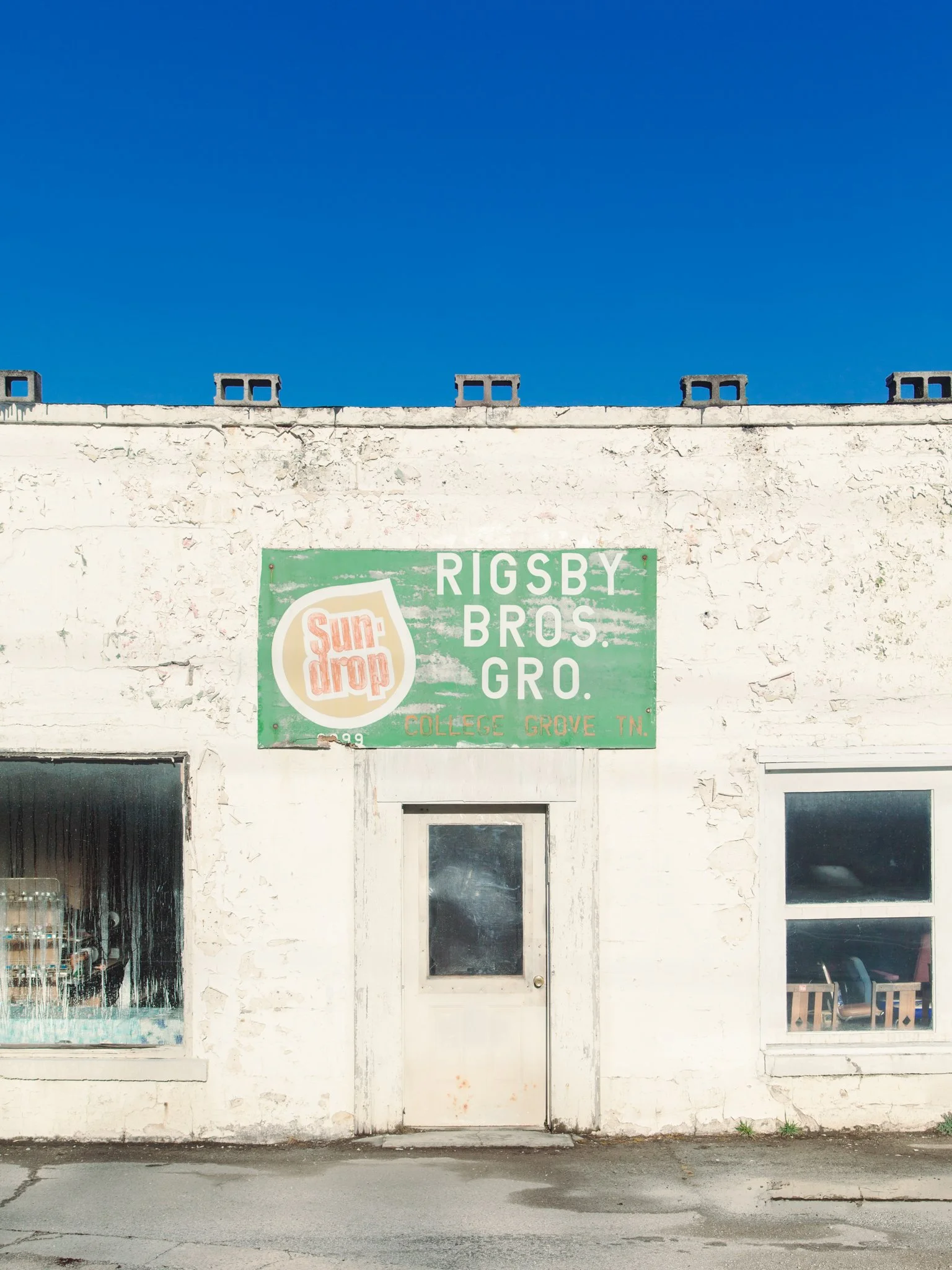 White weathered building with a door and two windows, a green sign with faded text and a logo, and a clear blue sky.