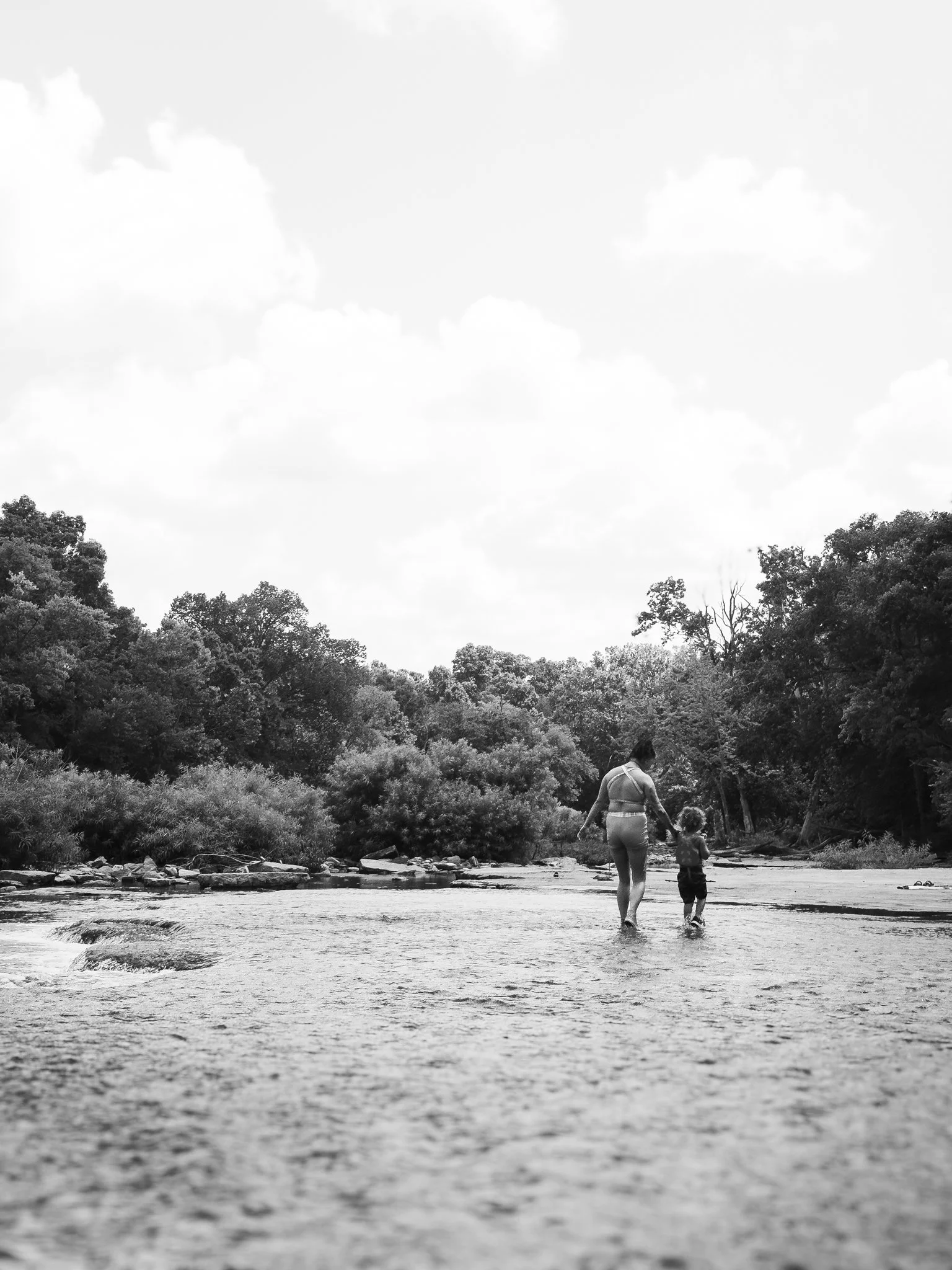 A woman and a young girl walking hand in hand along a riverbank with trees and a cloudy sky in the background.