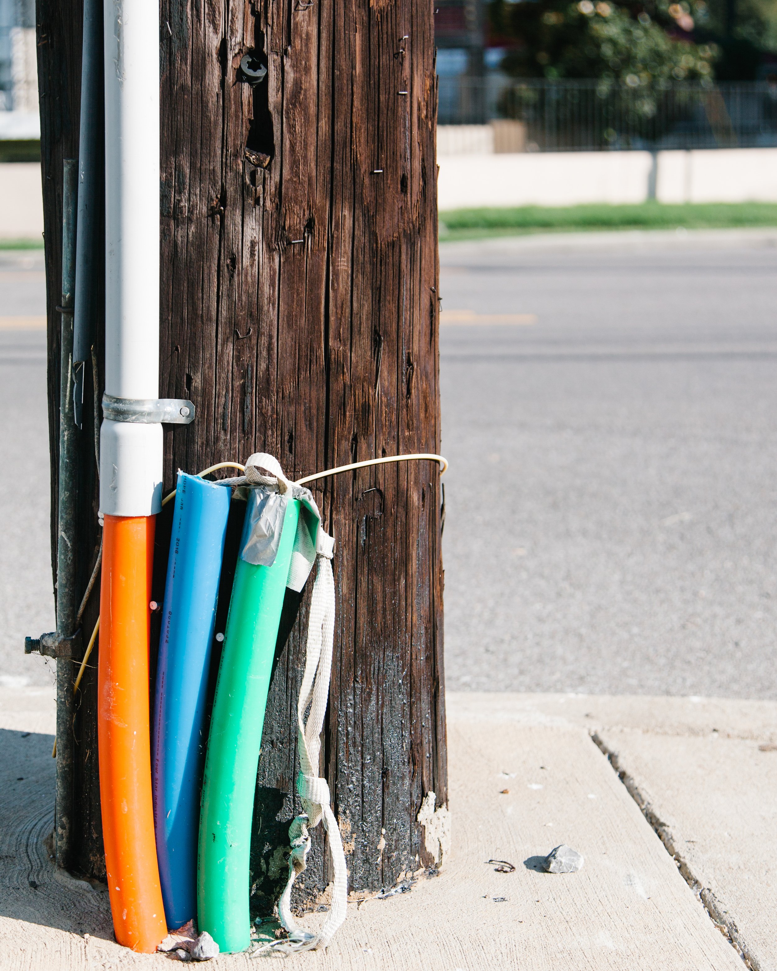 Colorful green, blue, and orange pipes connected and attached to a weathered wooden utility pole on a sidewalk.