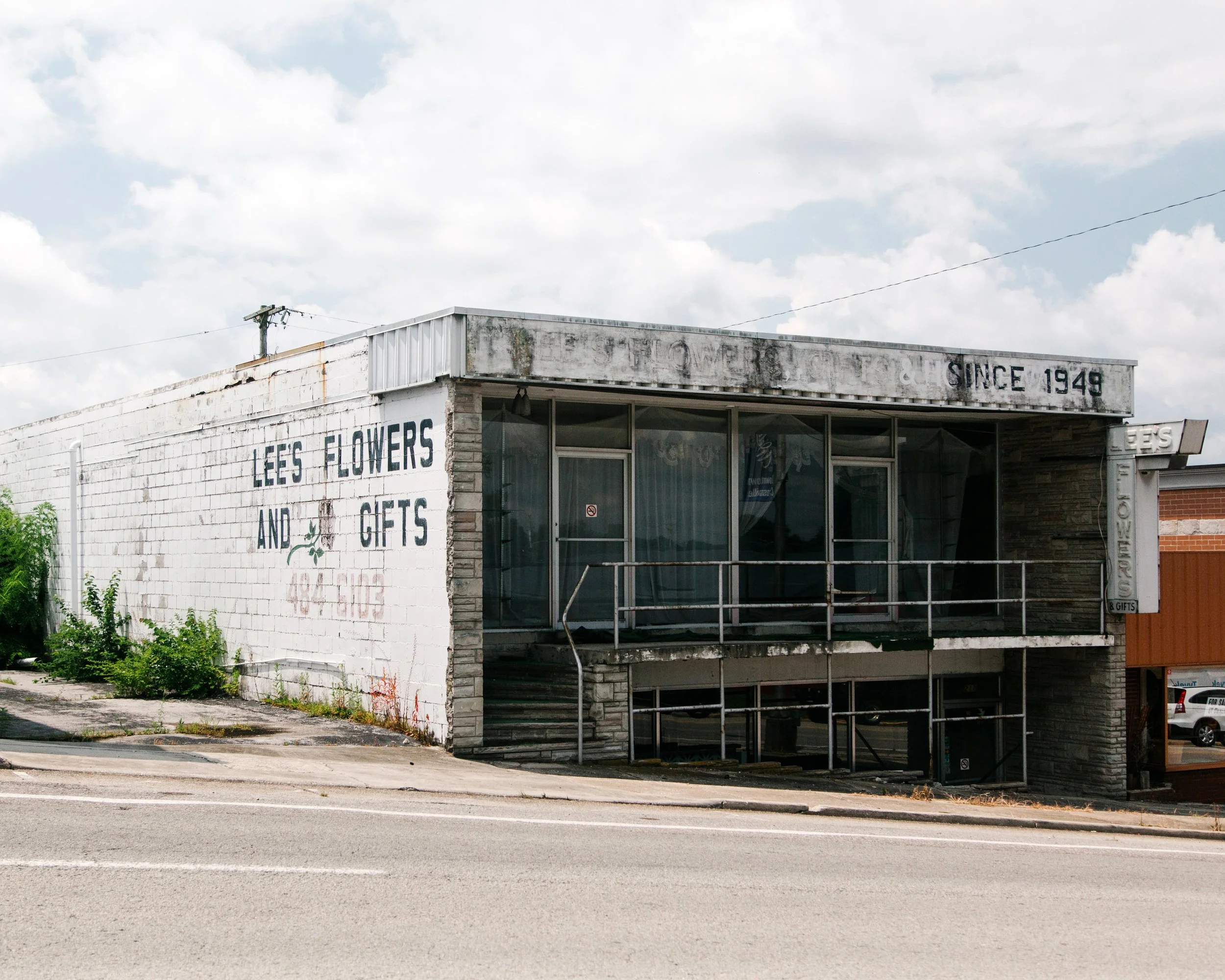Old white building with weathered sign that reads 'Since 1949' and another sign promoting flowers, gifts, and plants, with a large glass window at the corner and street in front under partly cloudy sky.
