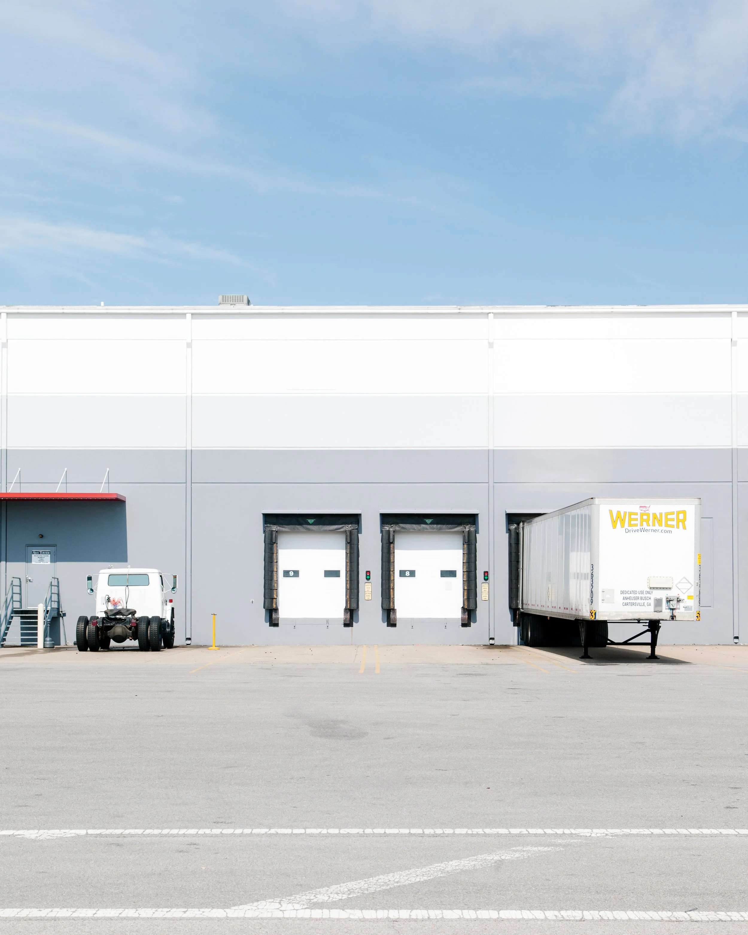 A parking lot in front of a large industrial building with three loading docks, a white delivery truck labeled 'WERNER,' and a small white van with equipment in the background. The sky is partly cloudy.