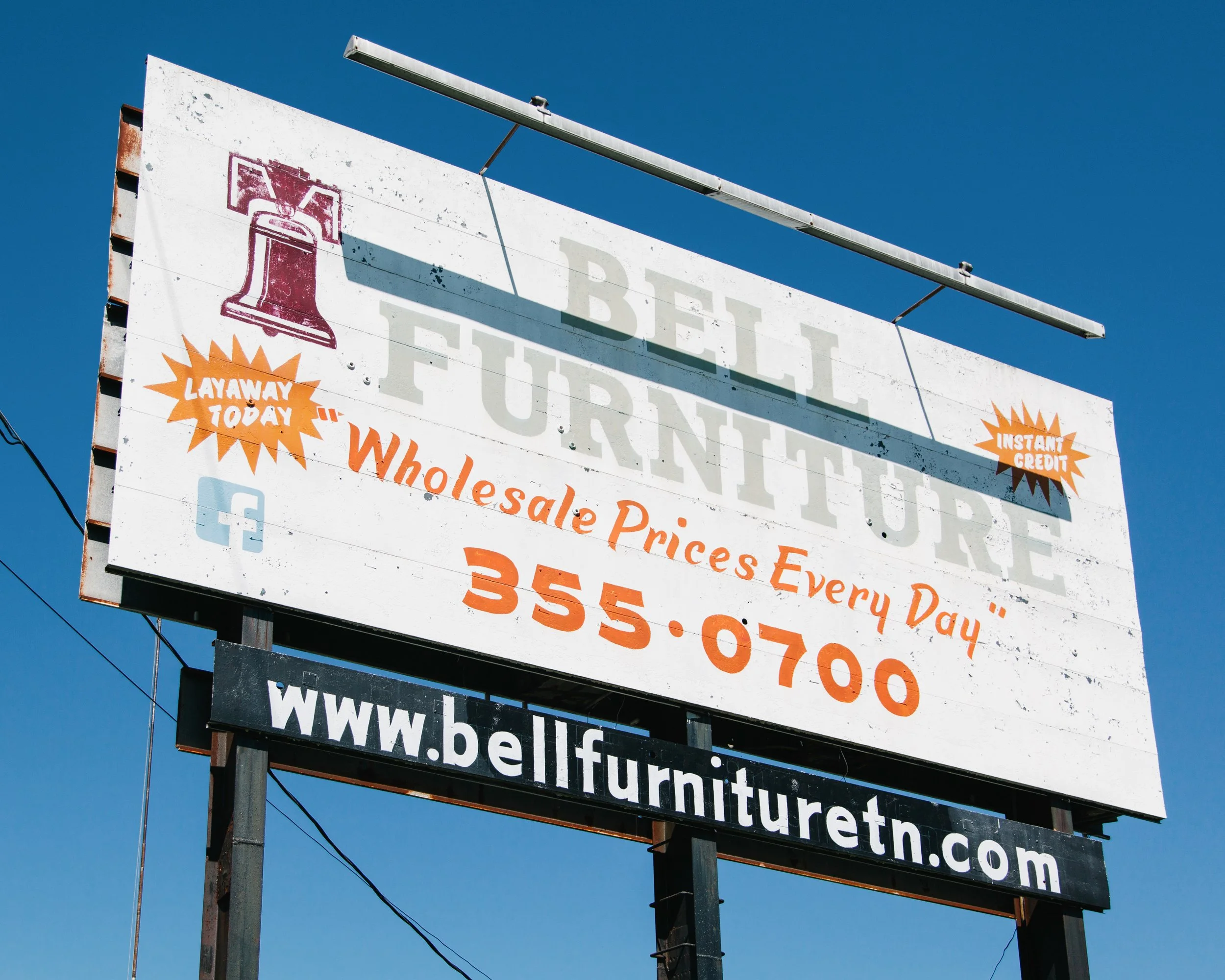 Billboard advertising wholesale prices with a red bell logo, orange starburst shapes with text, and a website URL. The billboard is mounted on a metal pole against a clear blue sky.