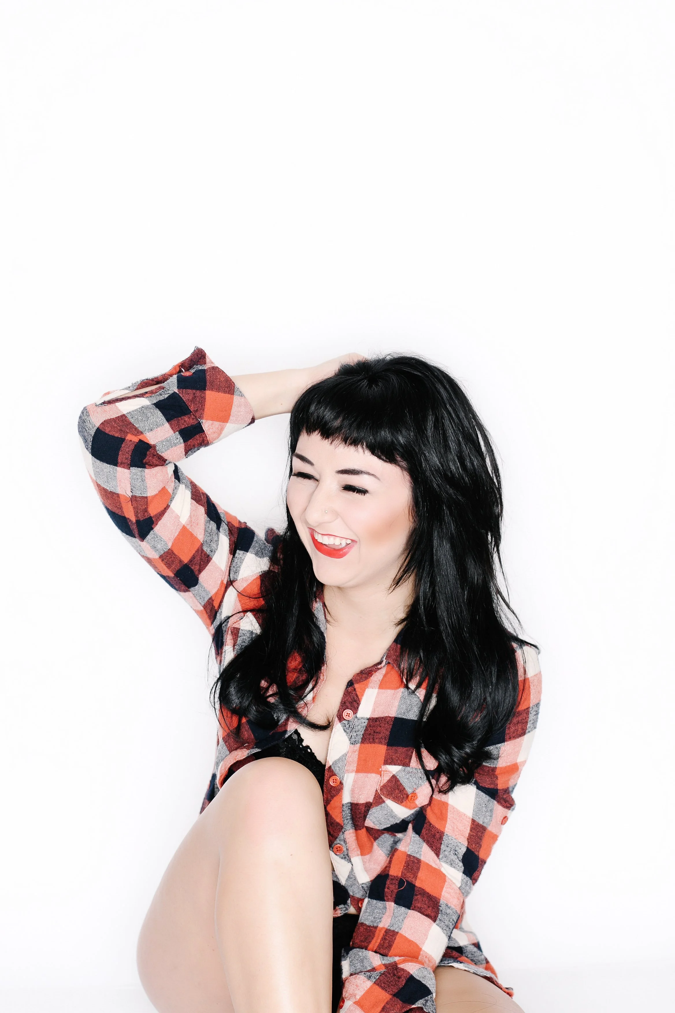 A woman with long black hair and a plaid shirt is smiling with her eyes closed against a white background. She has her left hand behind her head and her right arm is resting on her knee.