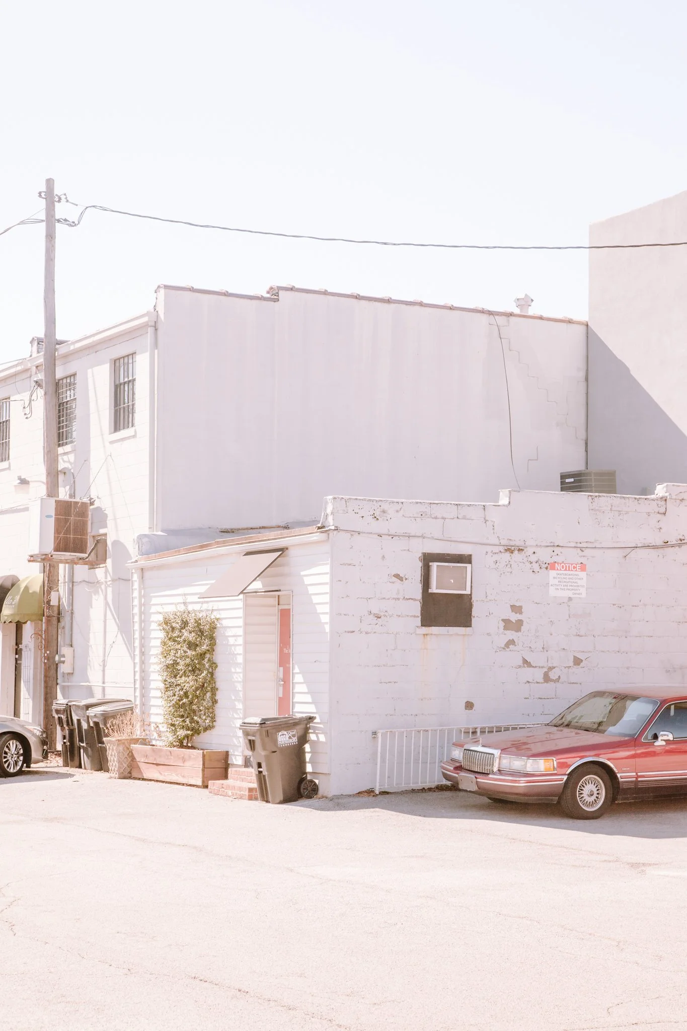 A white building with multiple windows and a small awning, parked cars, trash bins, and a tree outside, with utility poles and wires overhead, under a bright, clear sky.