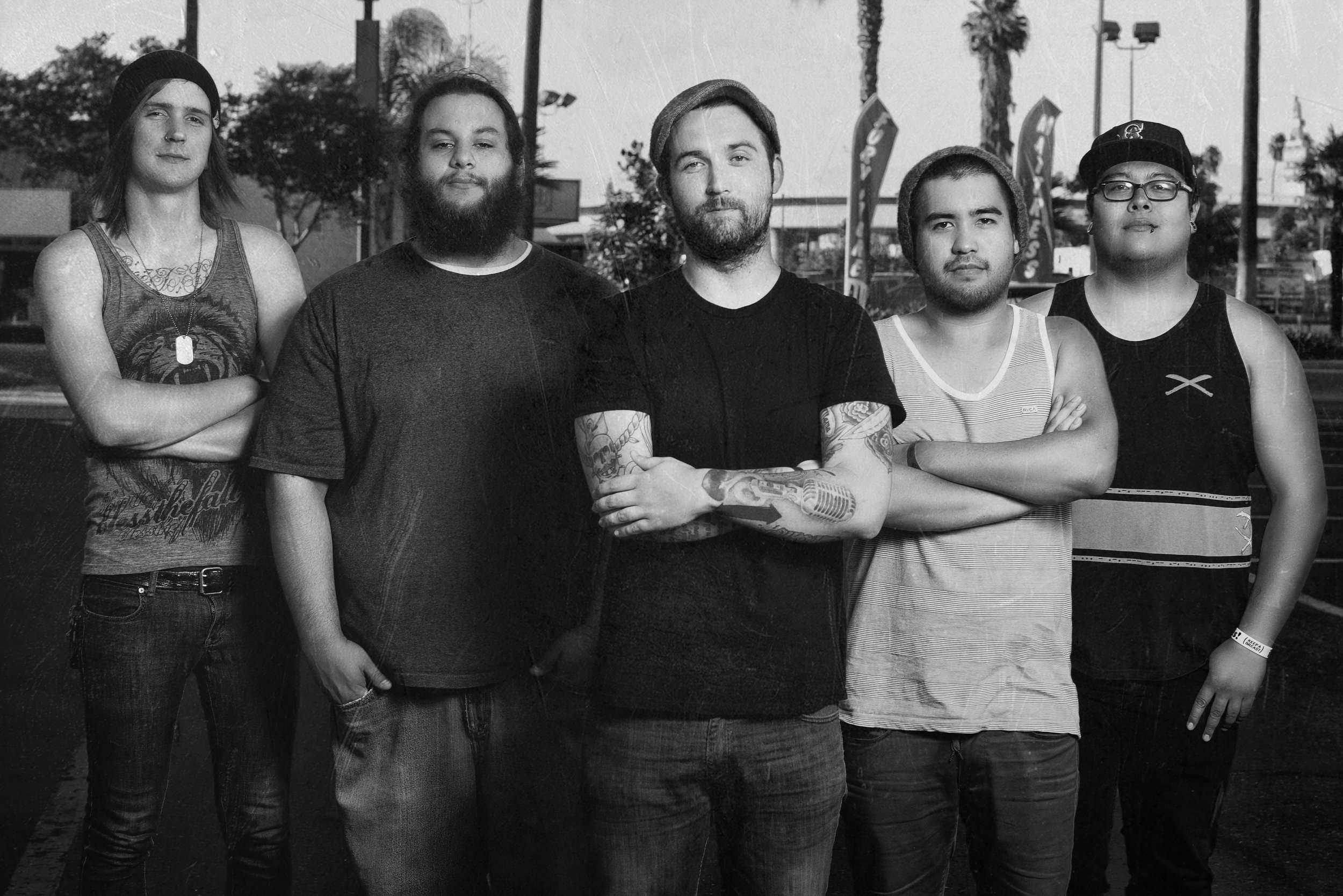 A group of five young men standing outdoors, with palm trees and flags in the background. They are wearing casual clothing and some have tattoos, beards, or glasses.
